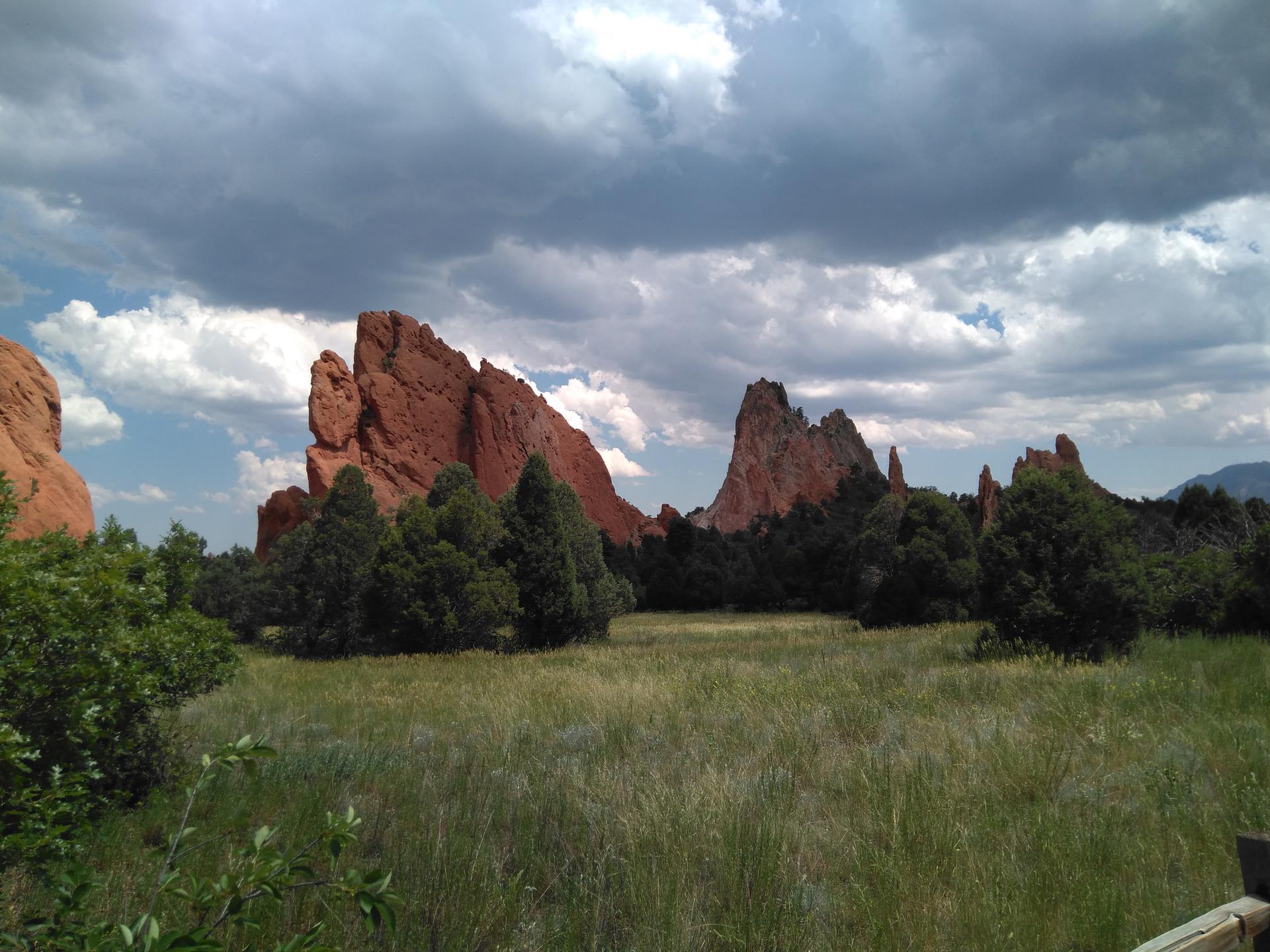 Majestic red rock formations rise above a green field under cloudy skies.
