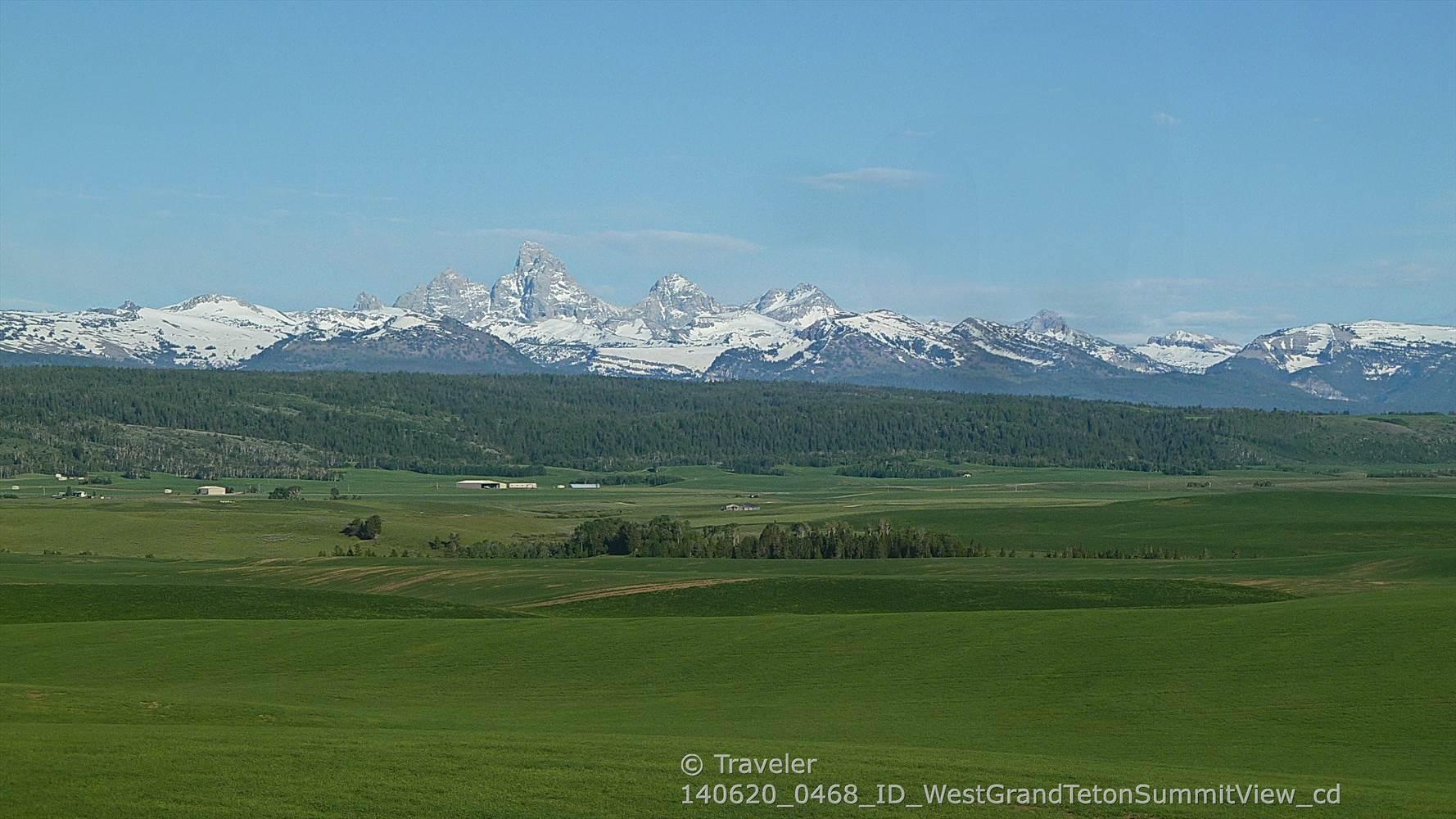 Snow-capped mountains overlook vast green fields and distant forests on a clear day.