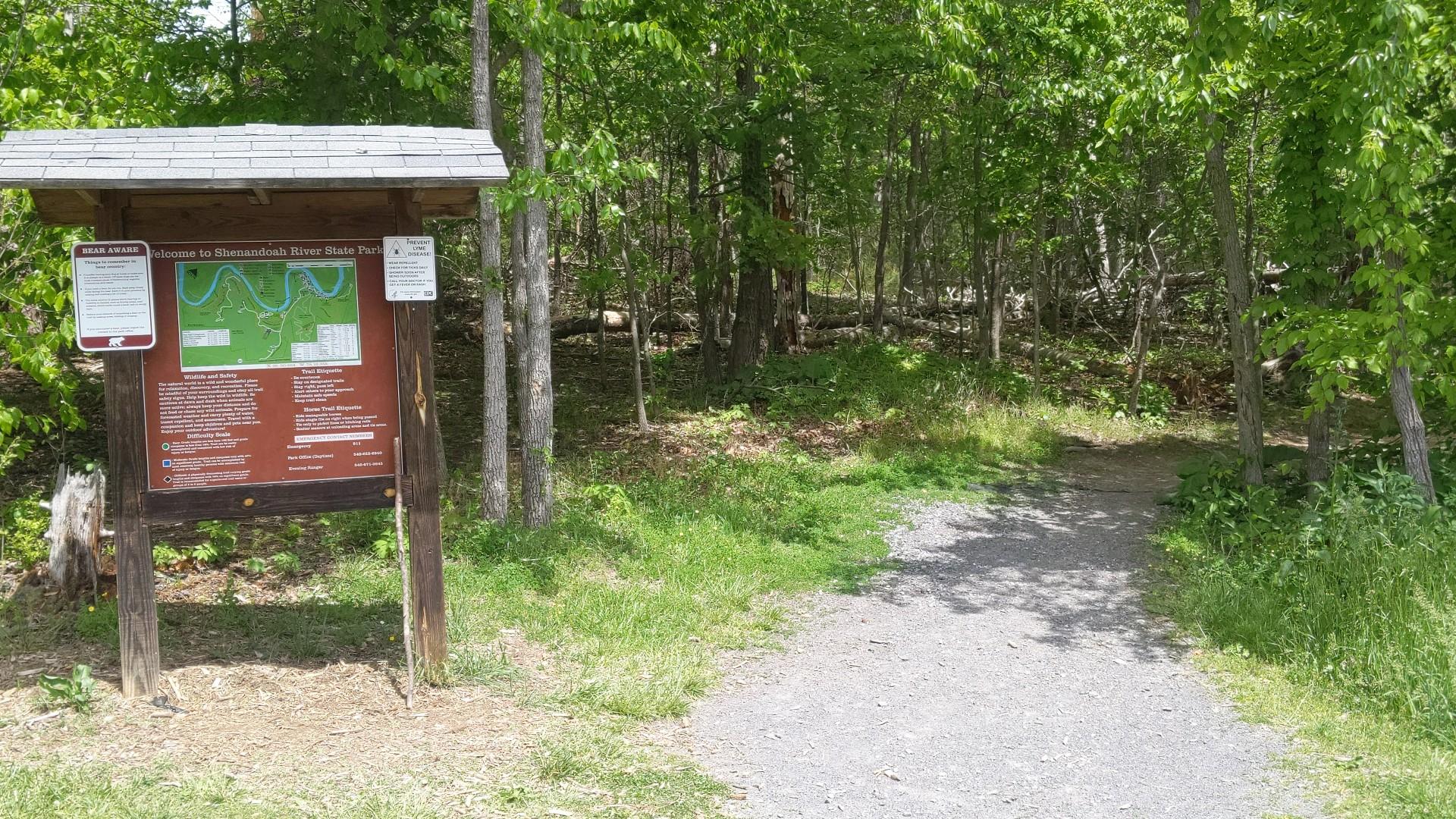 A trail information sign stands beside a gravel path entering a forest.