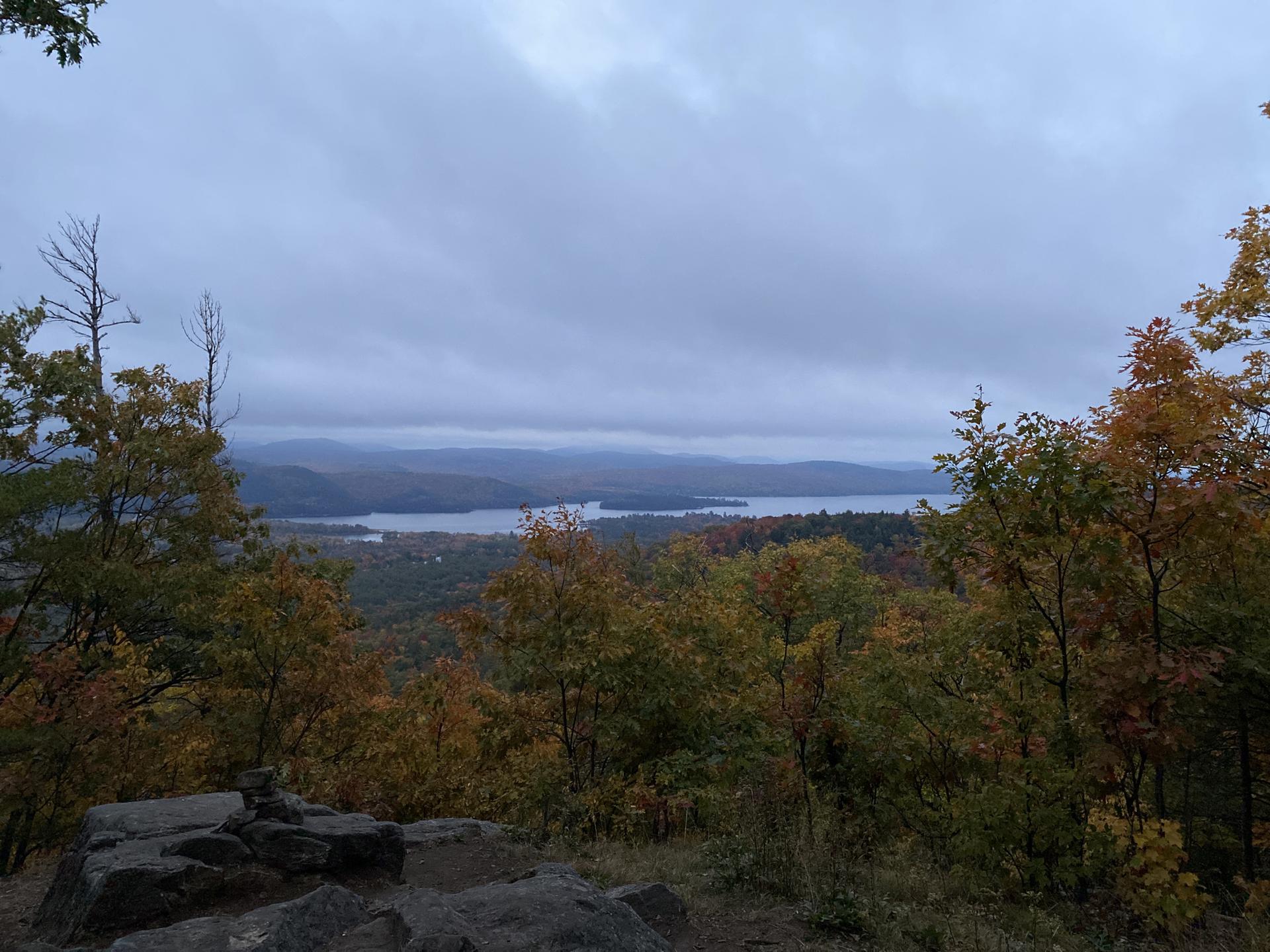 Overcast view of a lake surrounded by autumn trees and distant mountains.