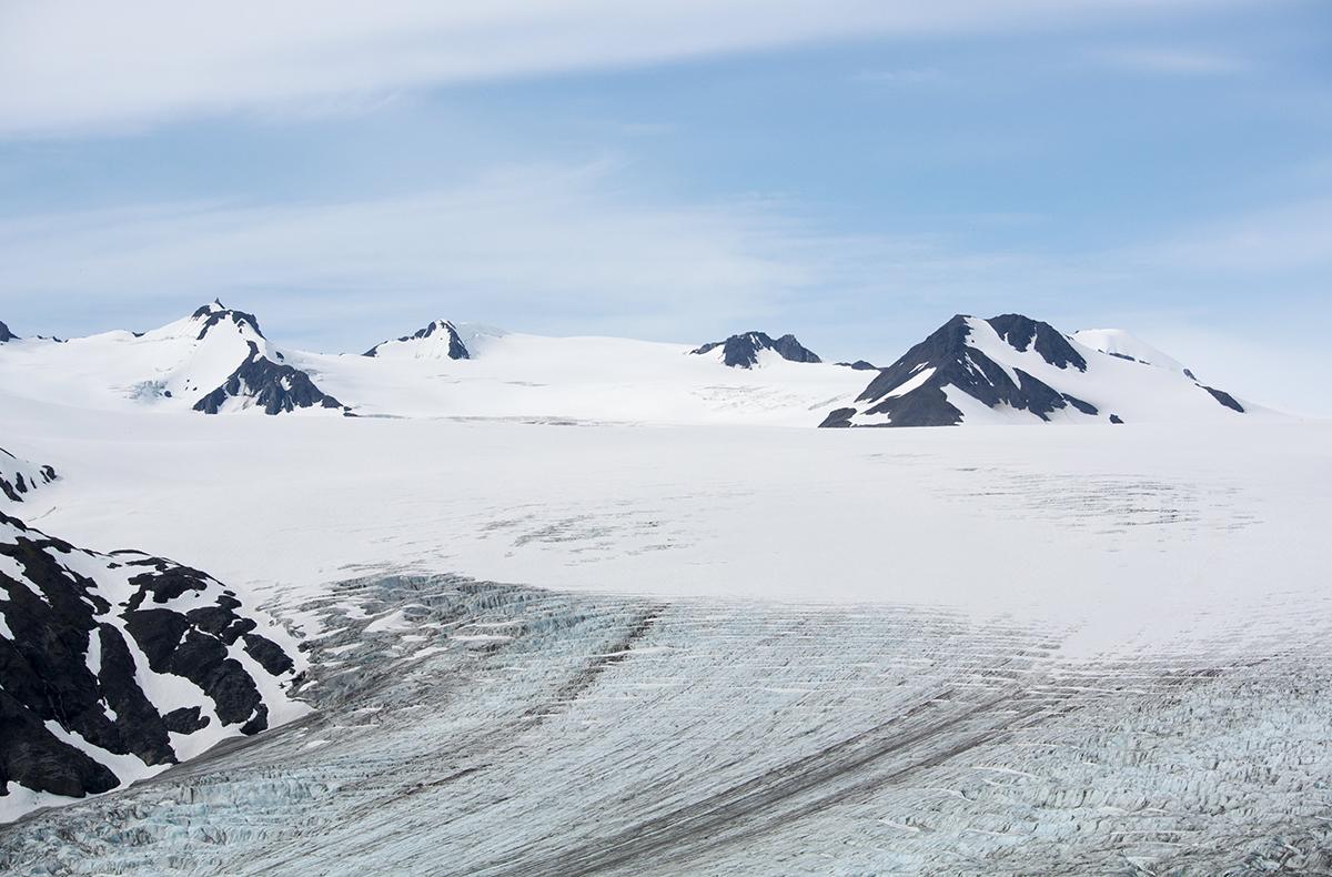 A vast, textured glacier surrounded by snow-covered peaks under a pale sky.
