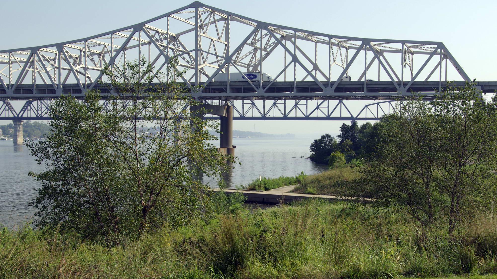 A large metal truss bridge spans a wide river with green banks.