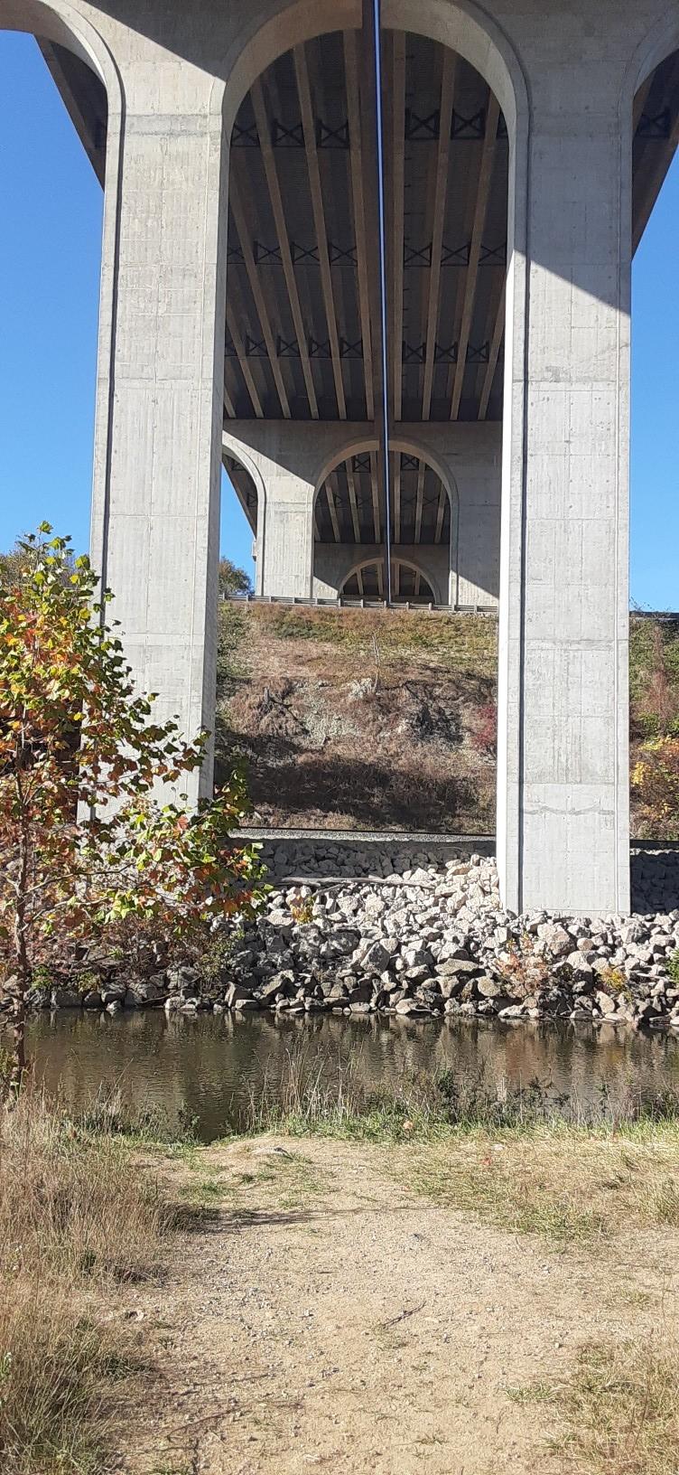 Concrete bridge arches over a river and dirt path on a sunny day.
