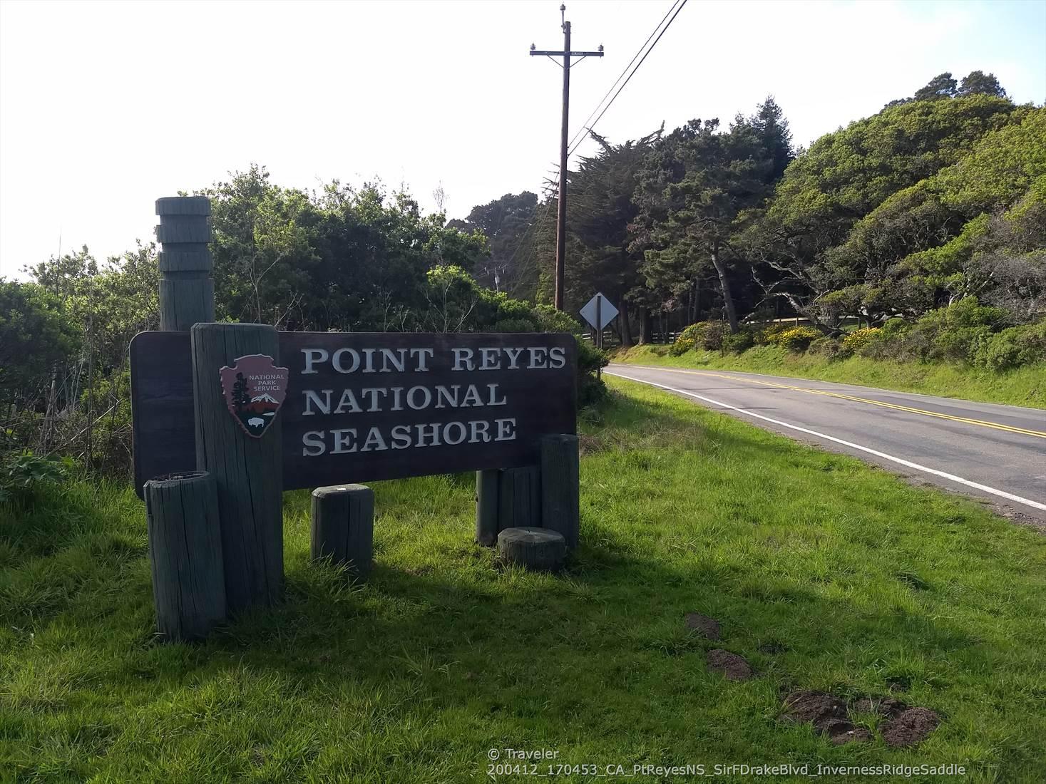 Large wooden sign by a paved road, surrounded by green trees and grass.