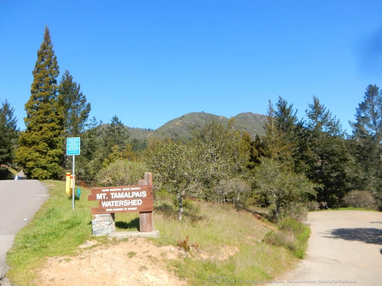 A wooden sign stands by a road, surrounded by trees and hills.