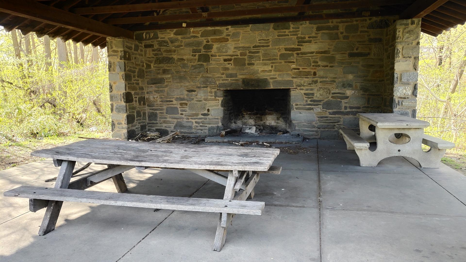 Wooden and concrete picnic tables inside a stone shelter with a fireplace.