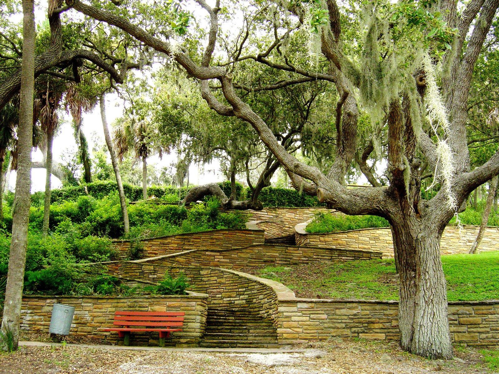 Terraced garden with stone walls, steps, and a red bench under large trees.