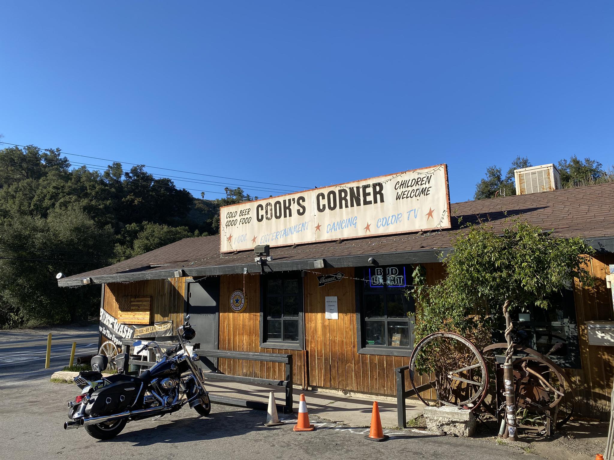 Rustic building with "Cook's Corner" sign, motorcycle, under clear blue sky.