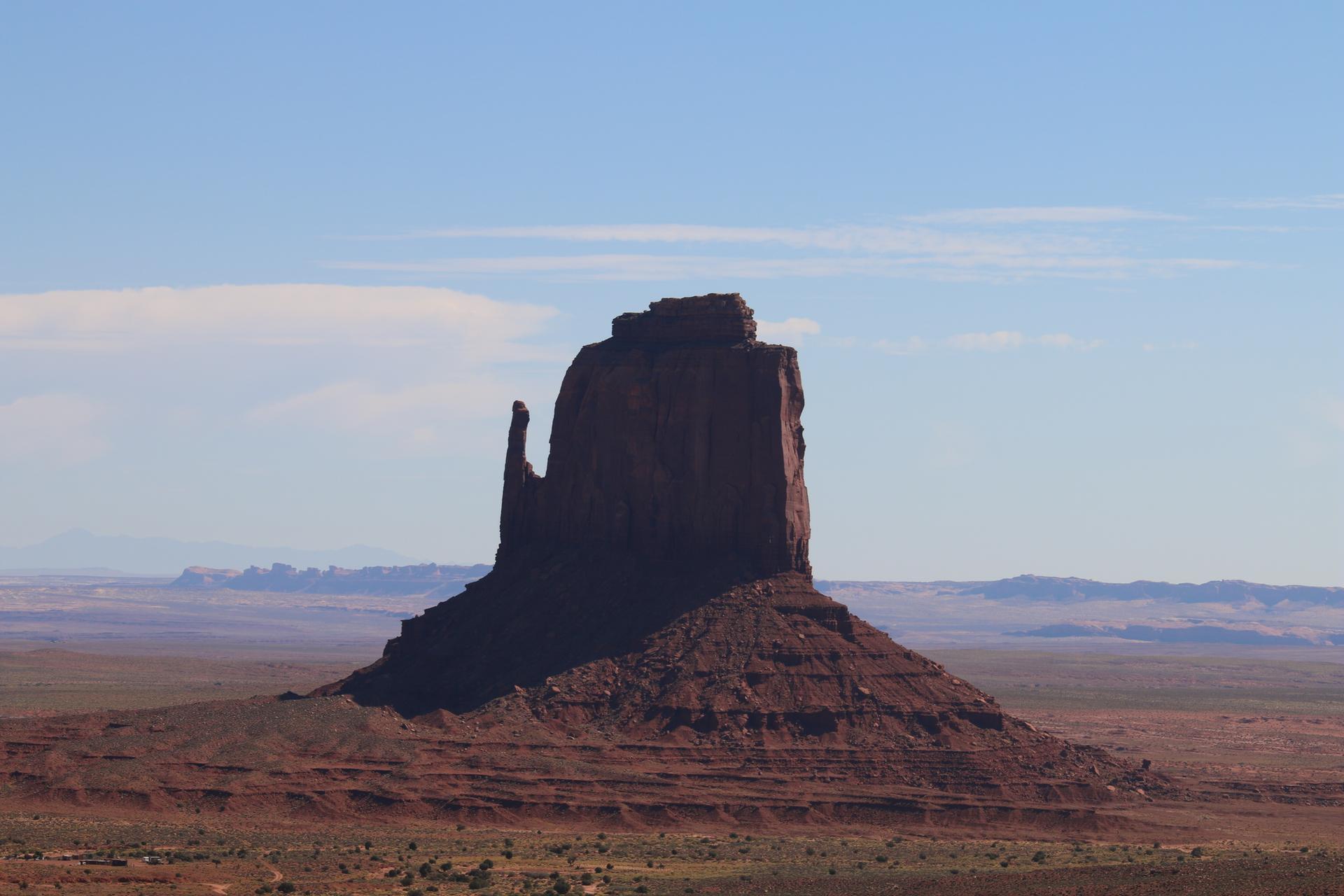 Distant red rock formation silhouetted against a light blue sky in a vast desert.