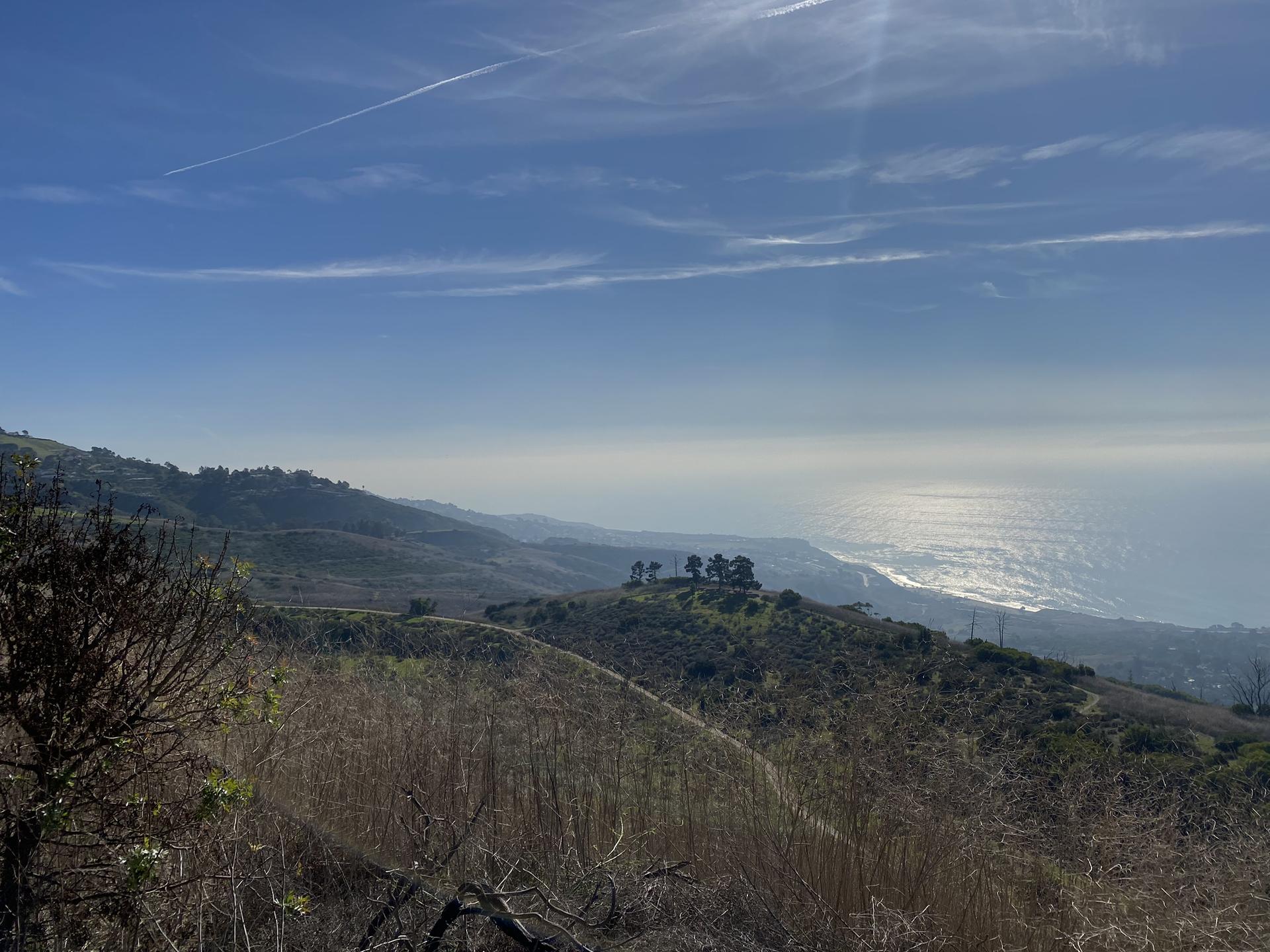 Sunny view of dry hills sloping towards a sparkling ocean under blue sky.