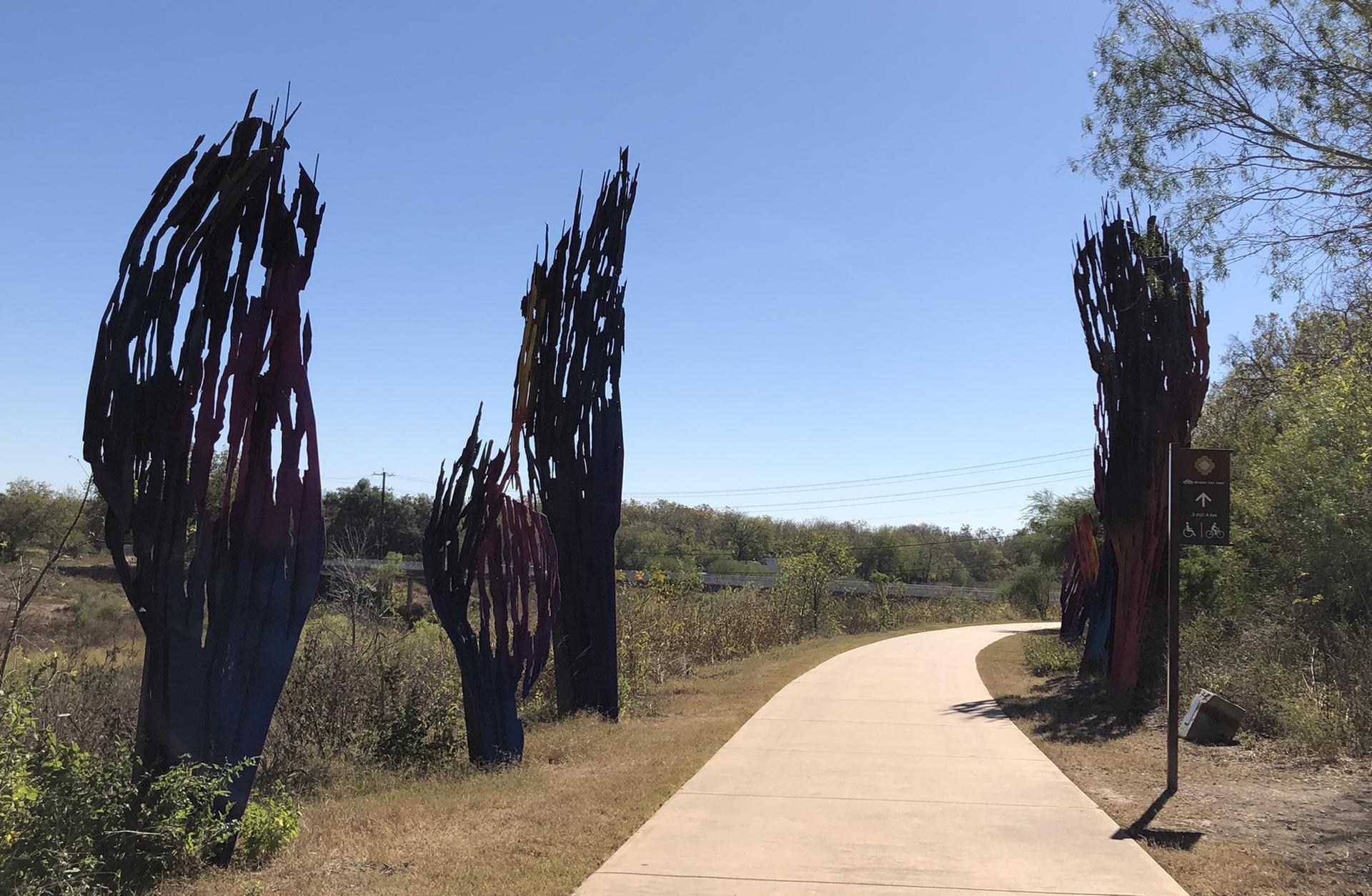 Paved path winding past tall, dark abstract sculptures under a clear blue sky.
