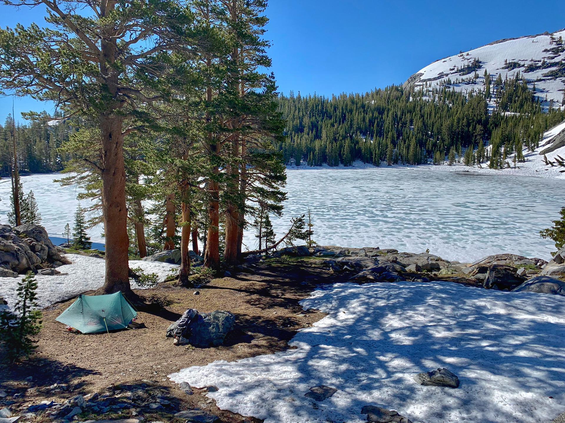 Green tent pitched near a partially frozen lake, surrounded by snow and pines.