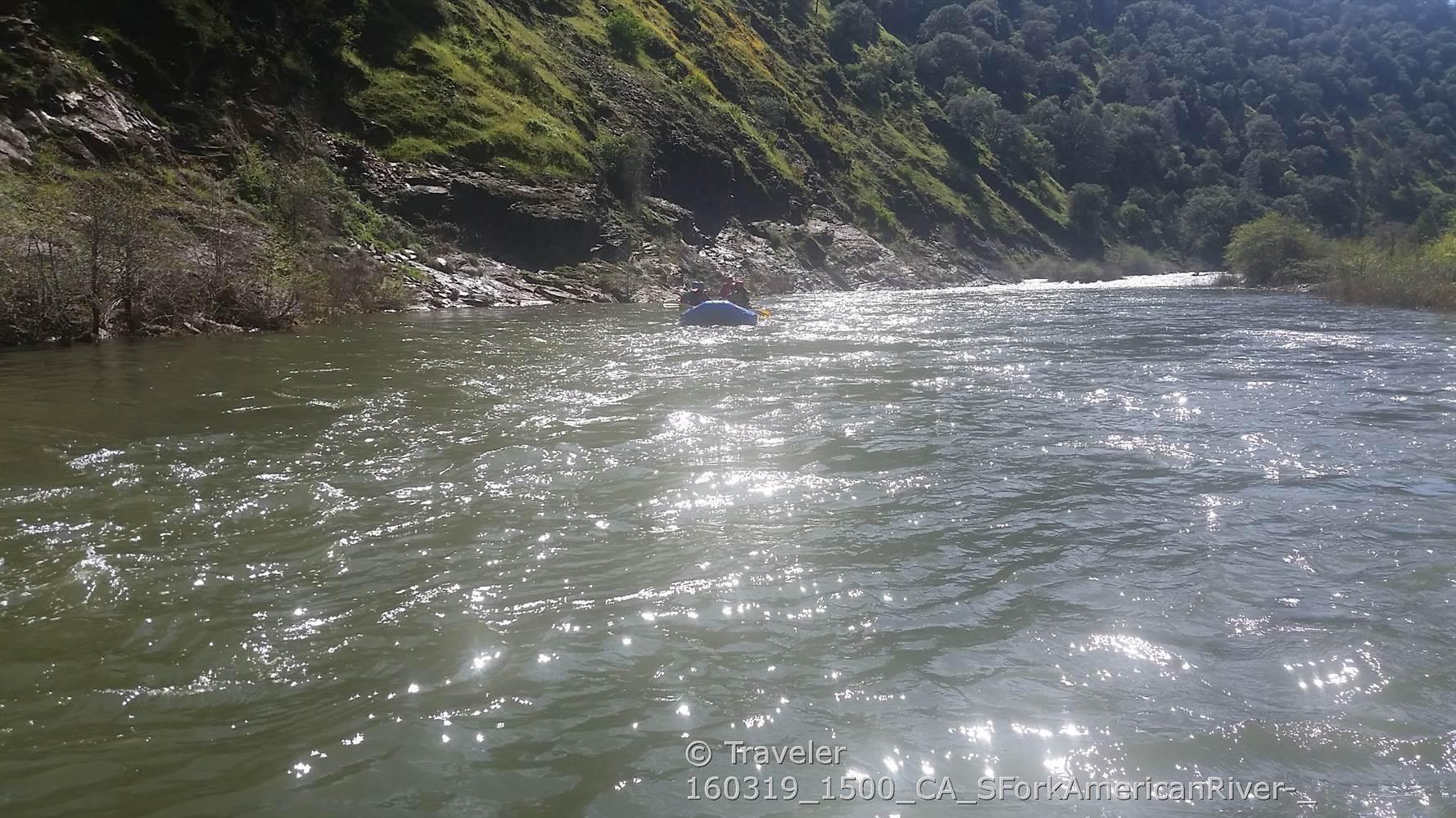 Person rafting on a sparkling river with steep green hillsides under bright sun.