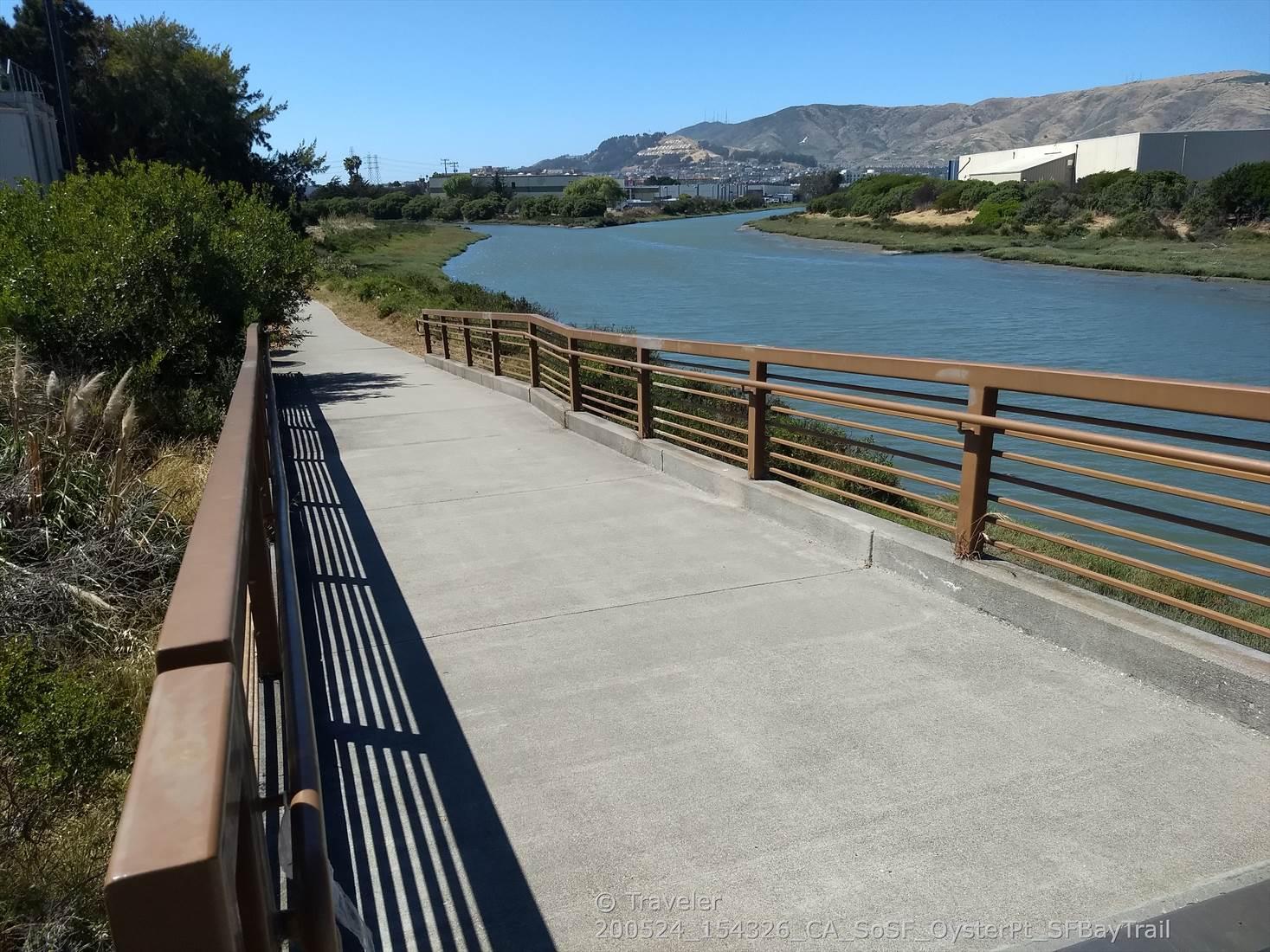 Paved walkway with railings next to a calm river, with hills in background.