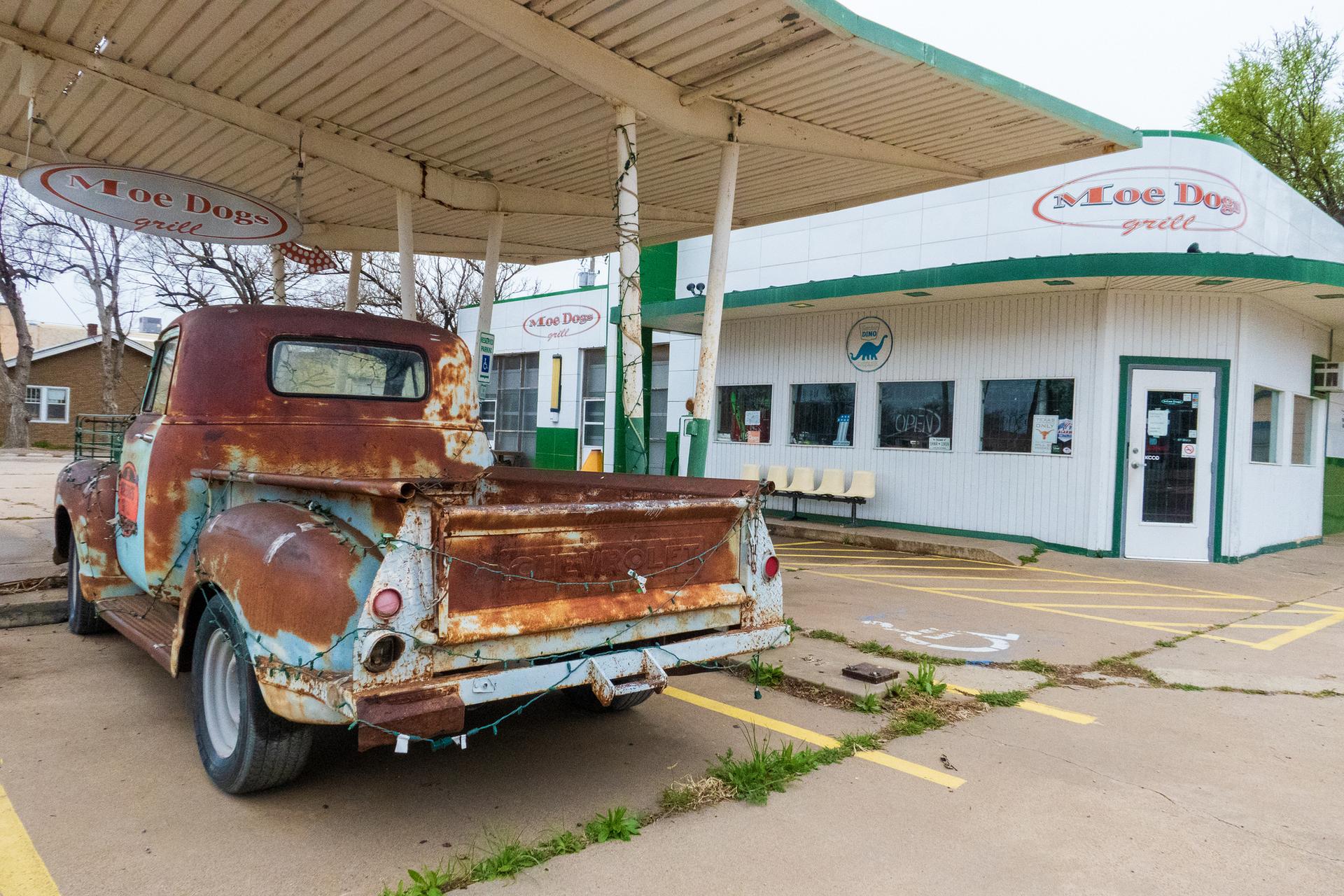 Rusty vintage pickup truck parked under a canopy next to a white building.