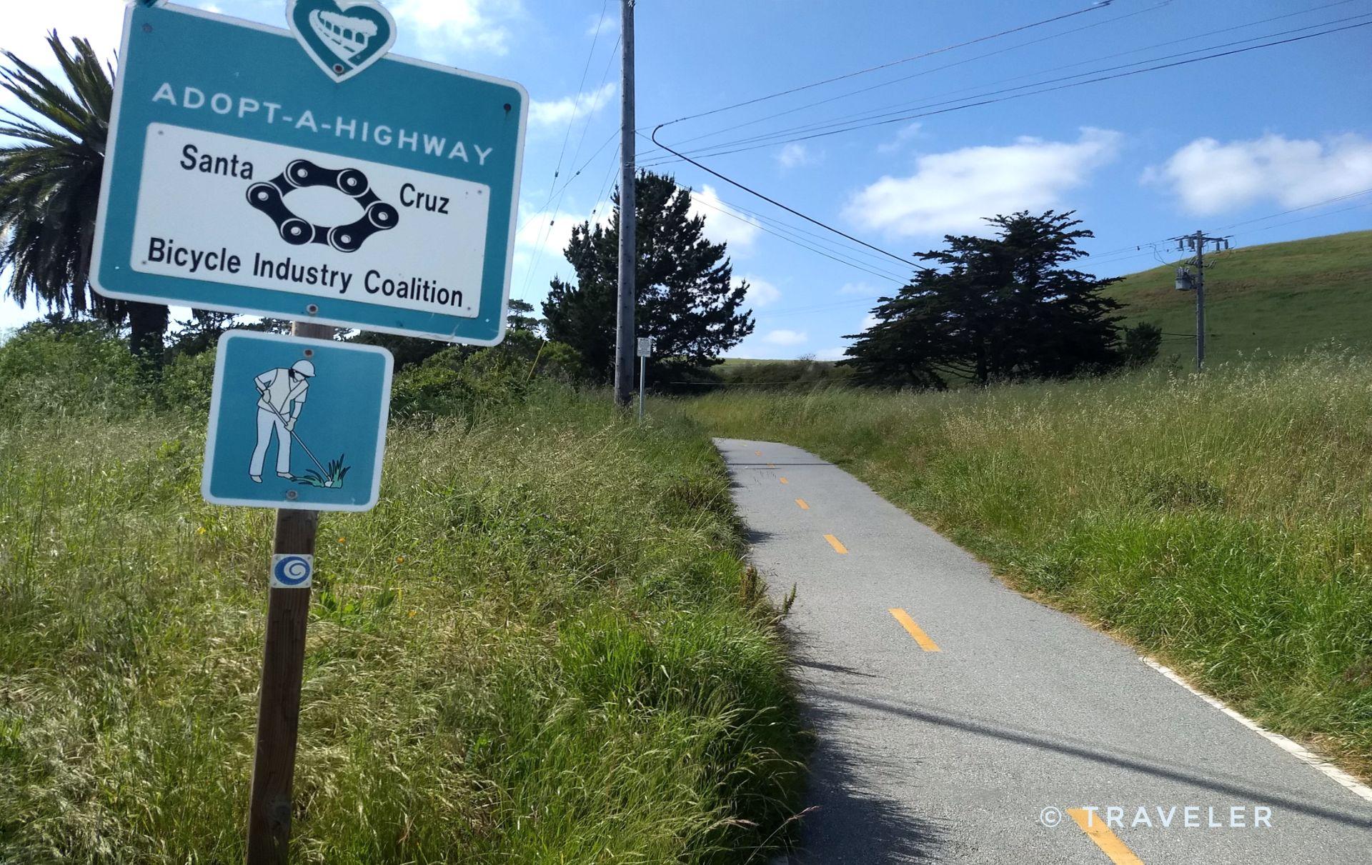 Road sign beside a paved path through grassy hills on a sunny day.