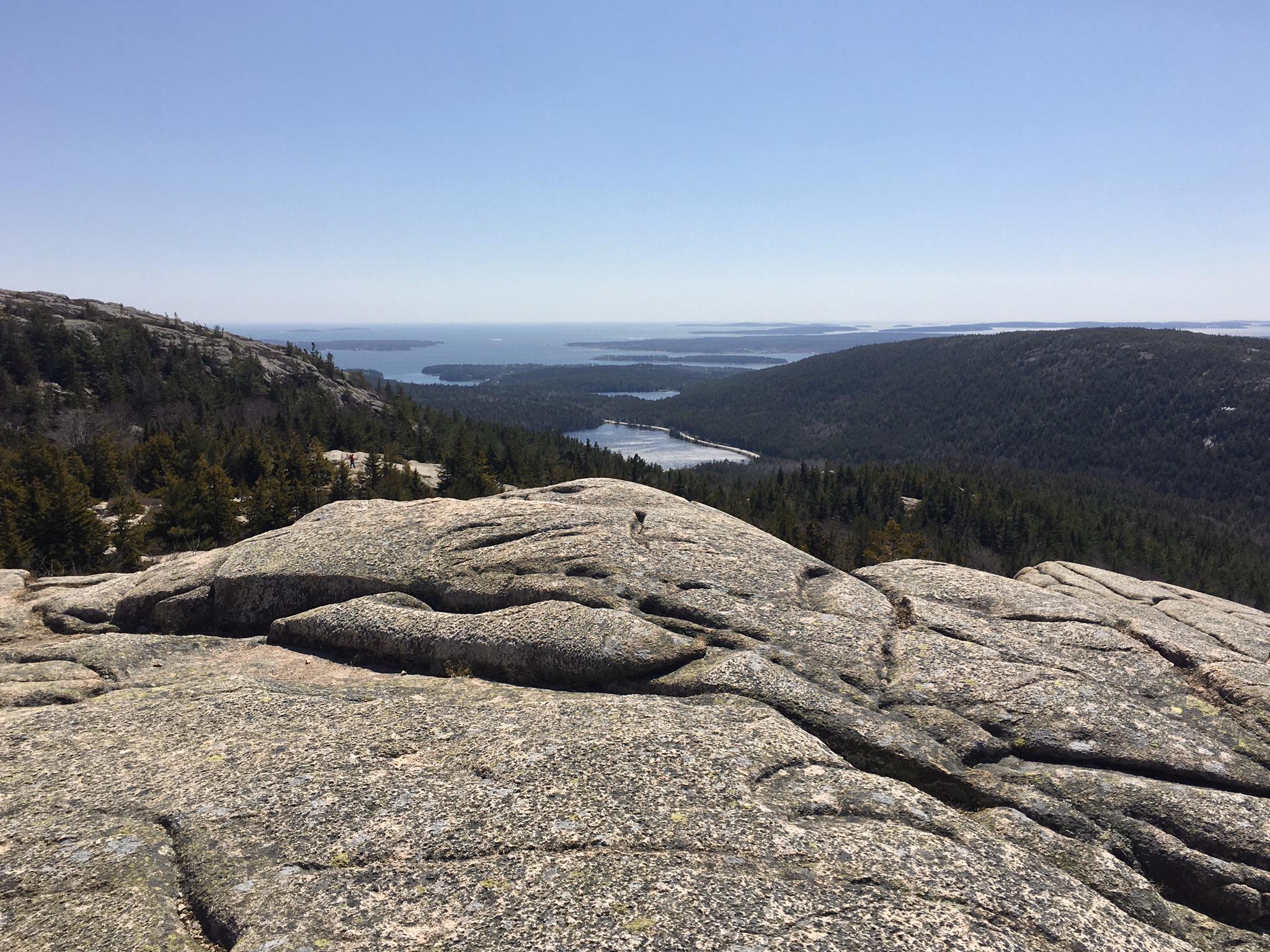 Large granite rock in foreground, overlooking forested valley, lake, and ocean.