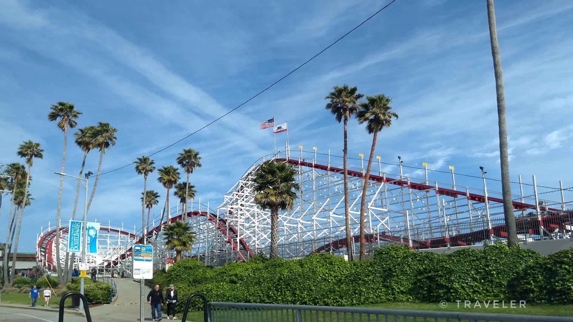 A large white and red roller coaster stands tall among palm trees.