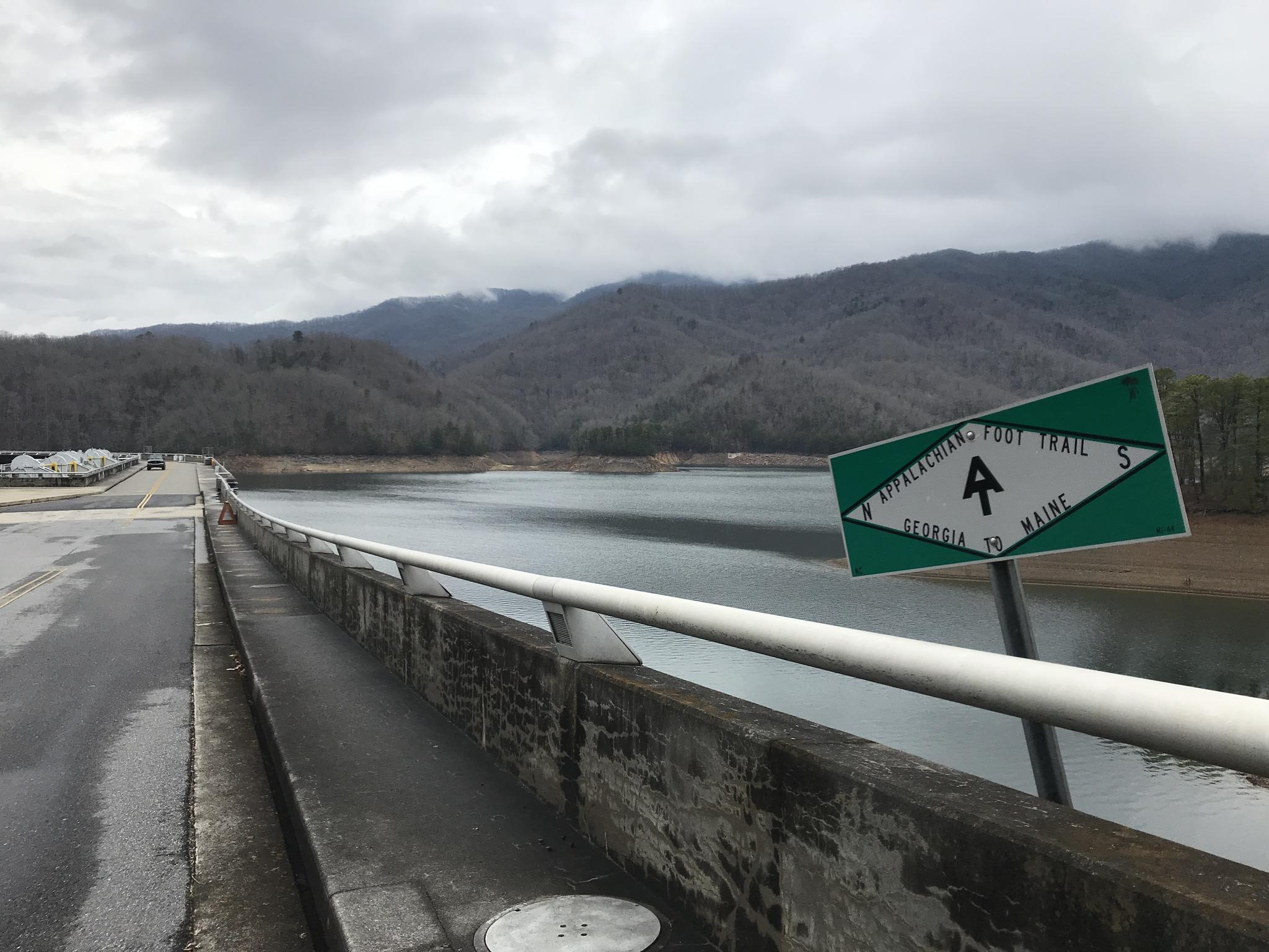 A bridge over a lake with a trail sign, mountains in the background on an overcast day.