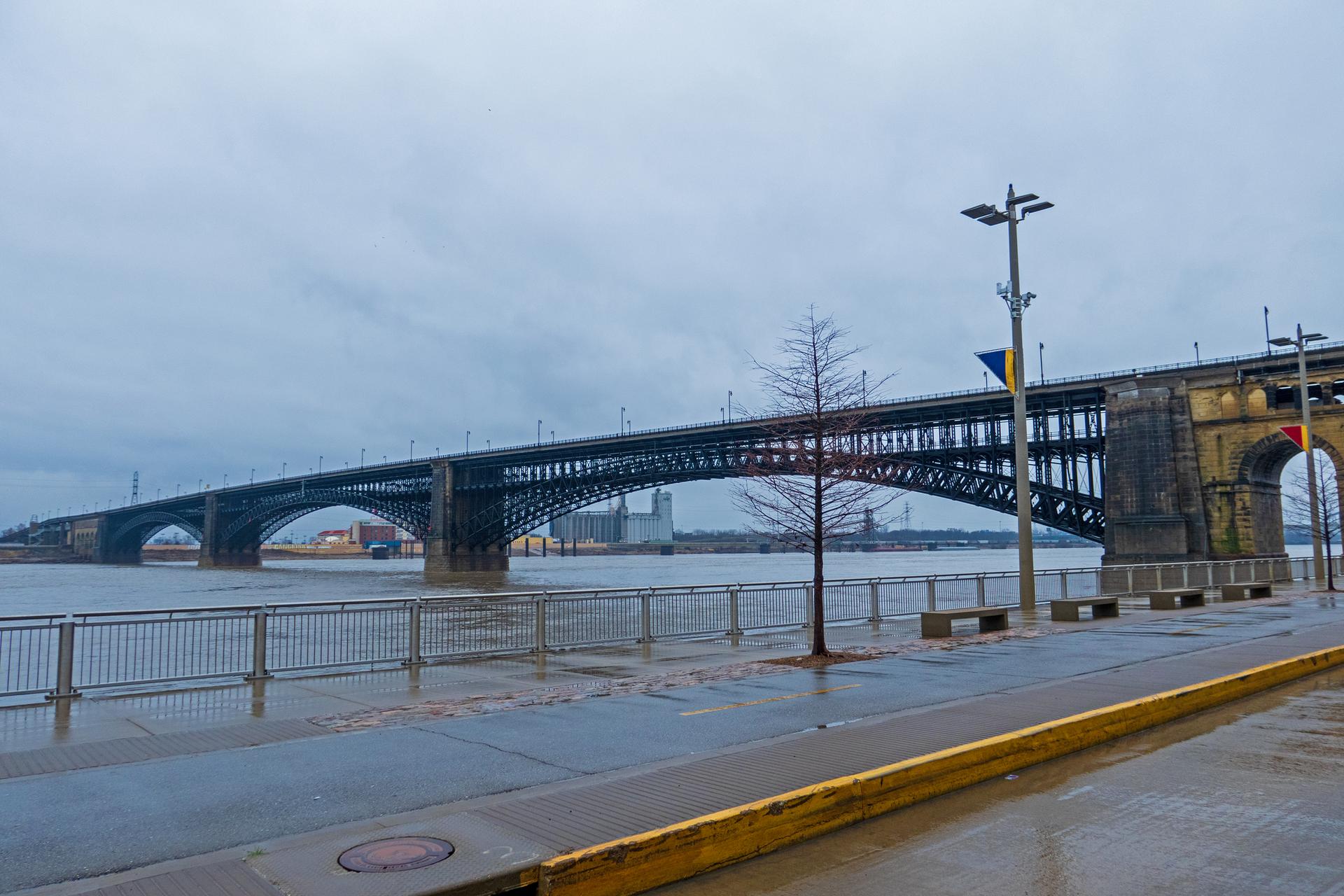 Wide metal bridge spans a river under a cloudy, grey sky.