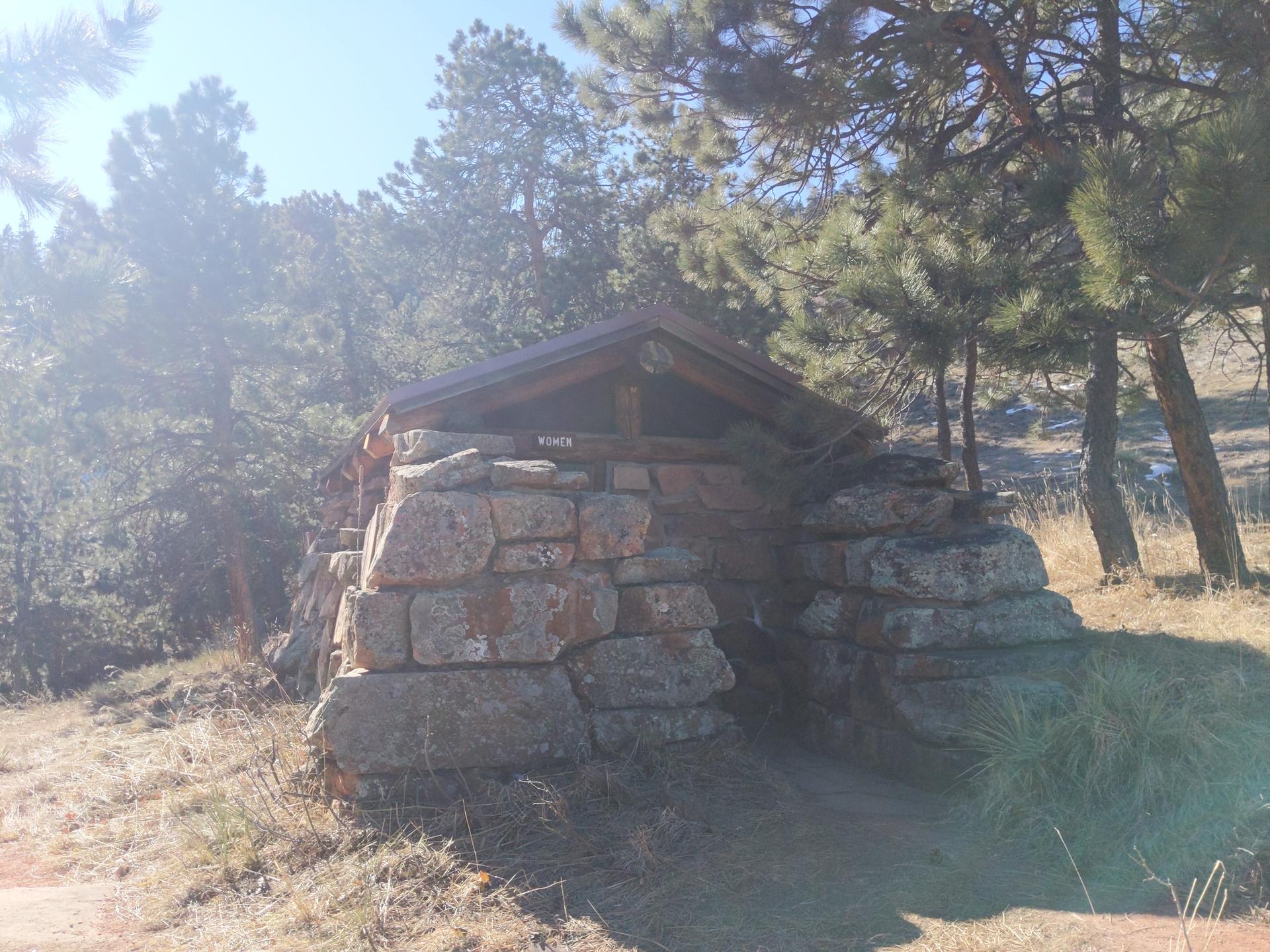 A stone shelter with a wooden roof nestled among pine trees on a sunny day.