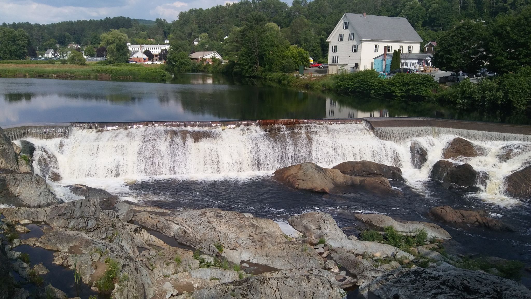 Wide waterfall cascading over rocks with buildings and calm river upstream. - Cross New Hampshire Adventure Trail