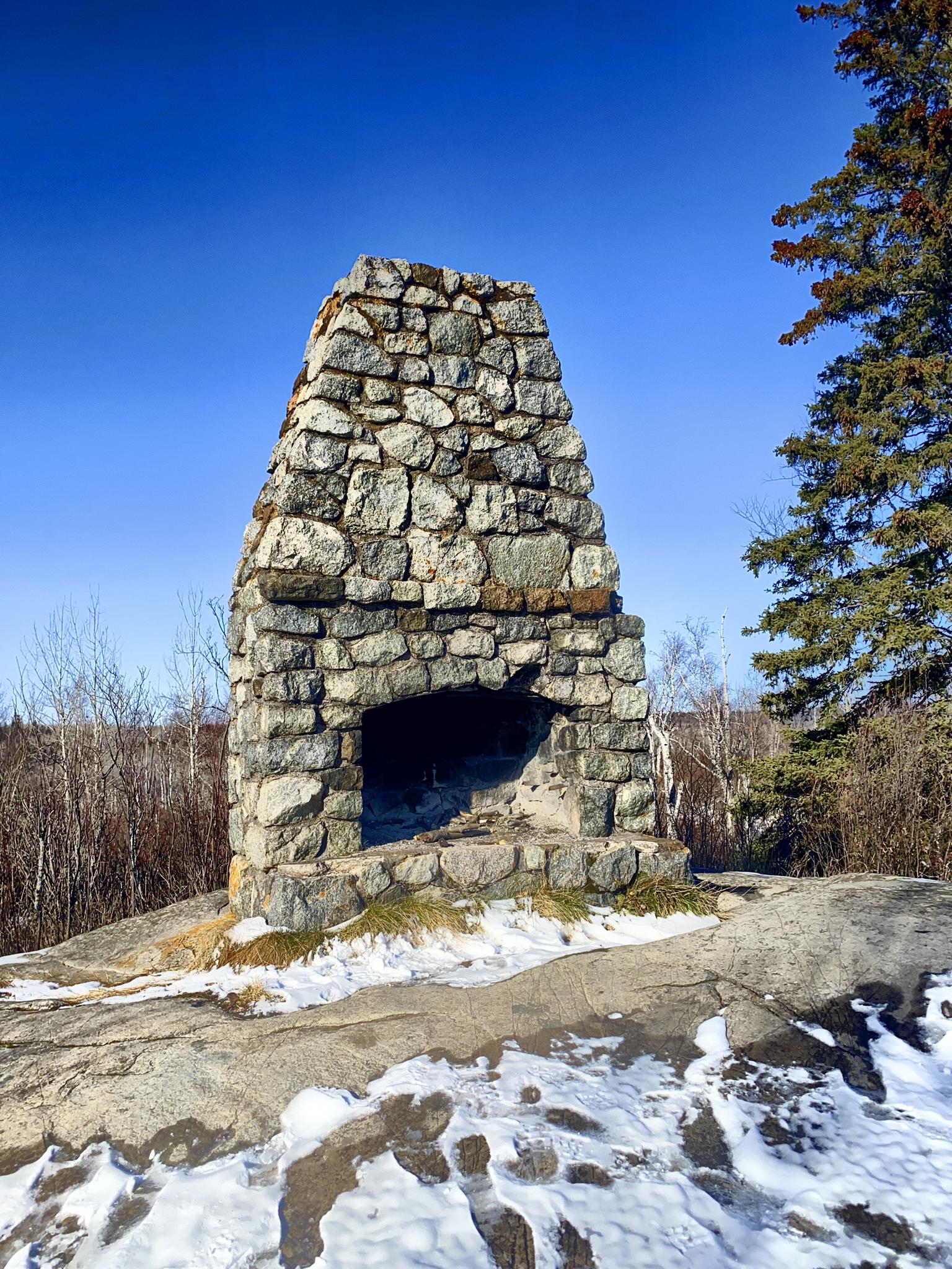 A large stone outdoor fireplace on snowy ground under a clear blue sky.
