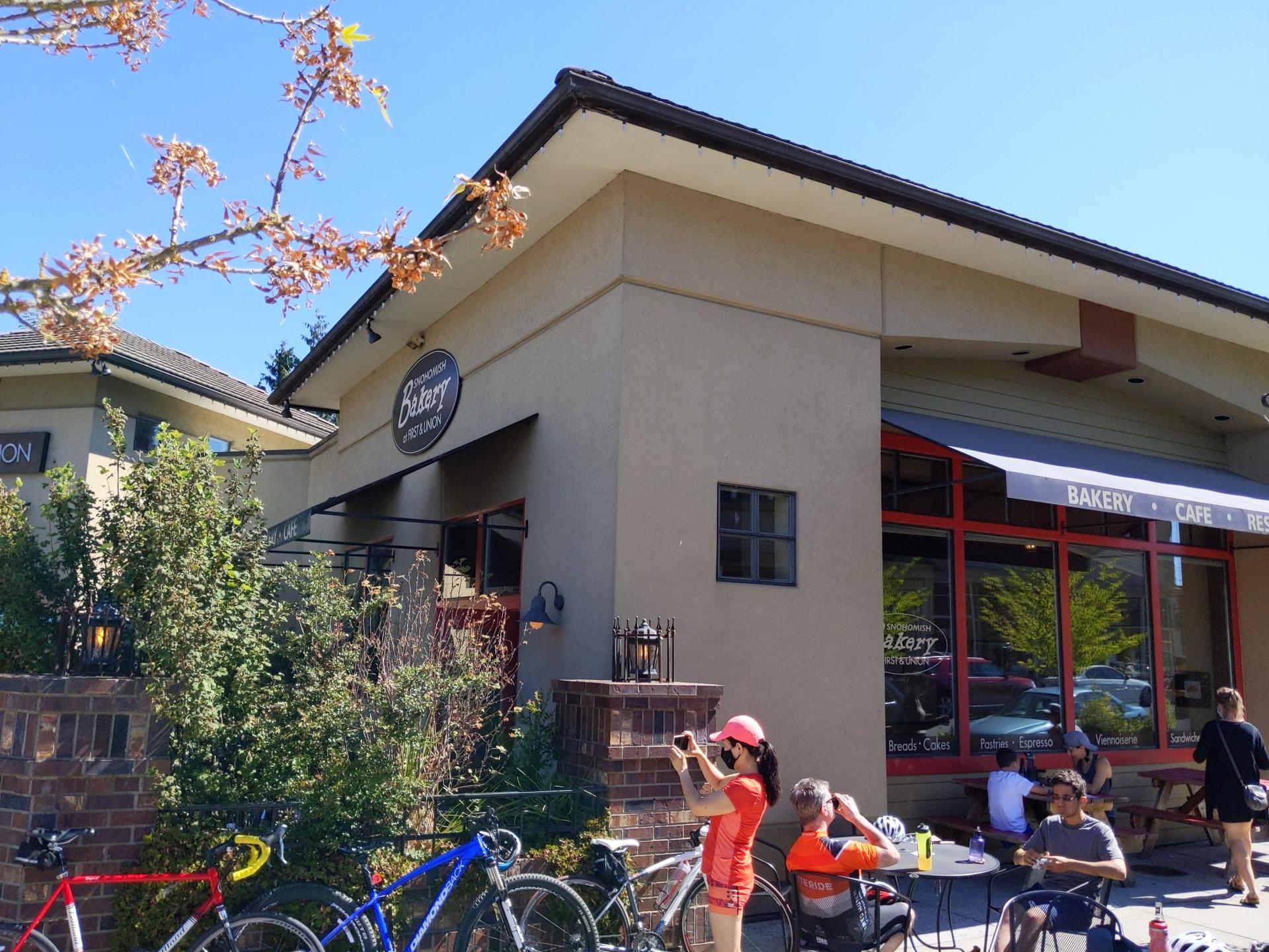 People relaxing outside a sunny bakery cafe with bicycles parked nearby.