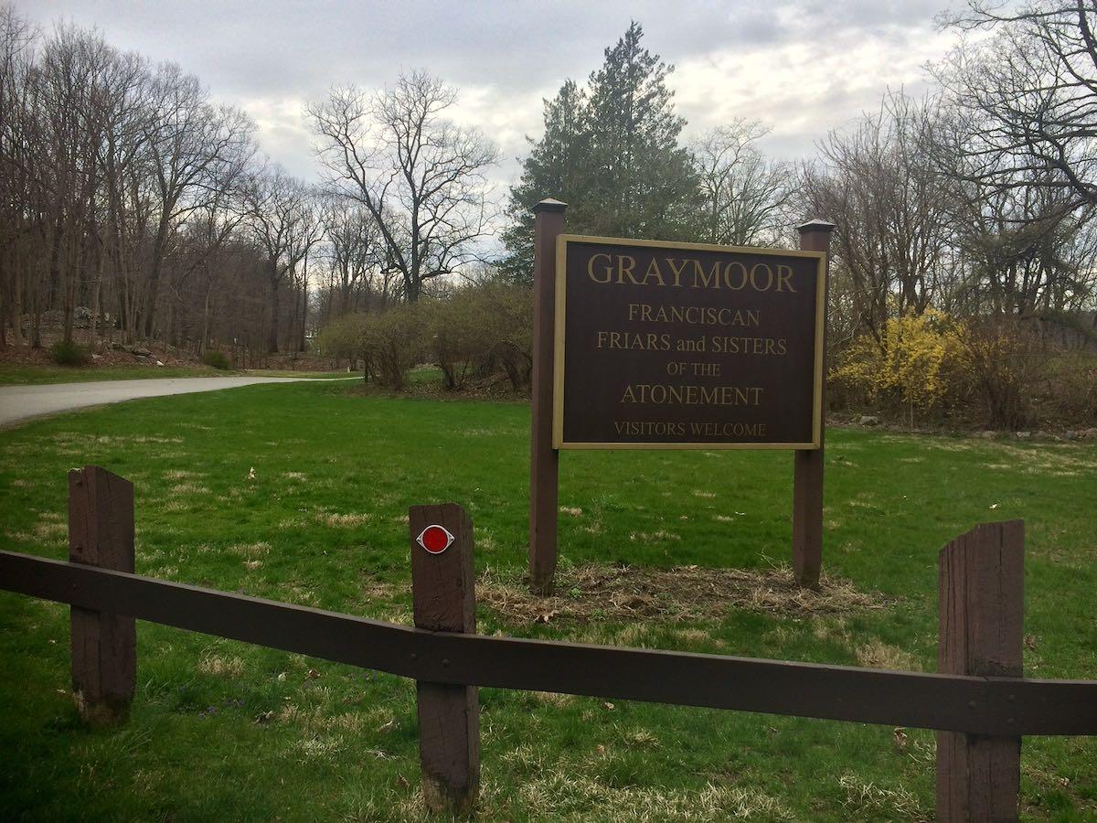 A brown sign stands beside a paved path and grassy area under an overcast sky.