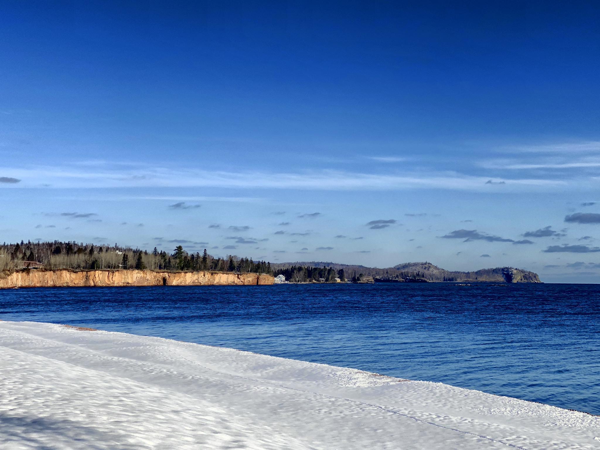 A snowy foreground overlooks deep blue water and distant forested cliffs under a clear sky.