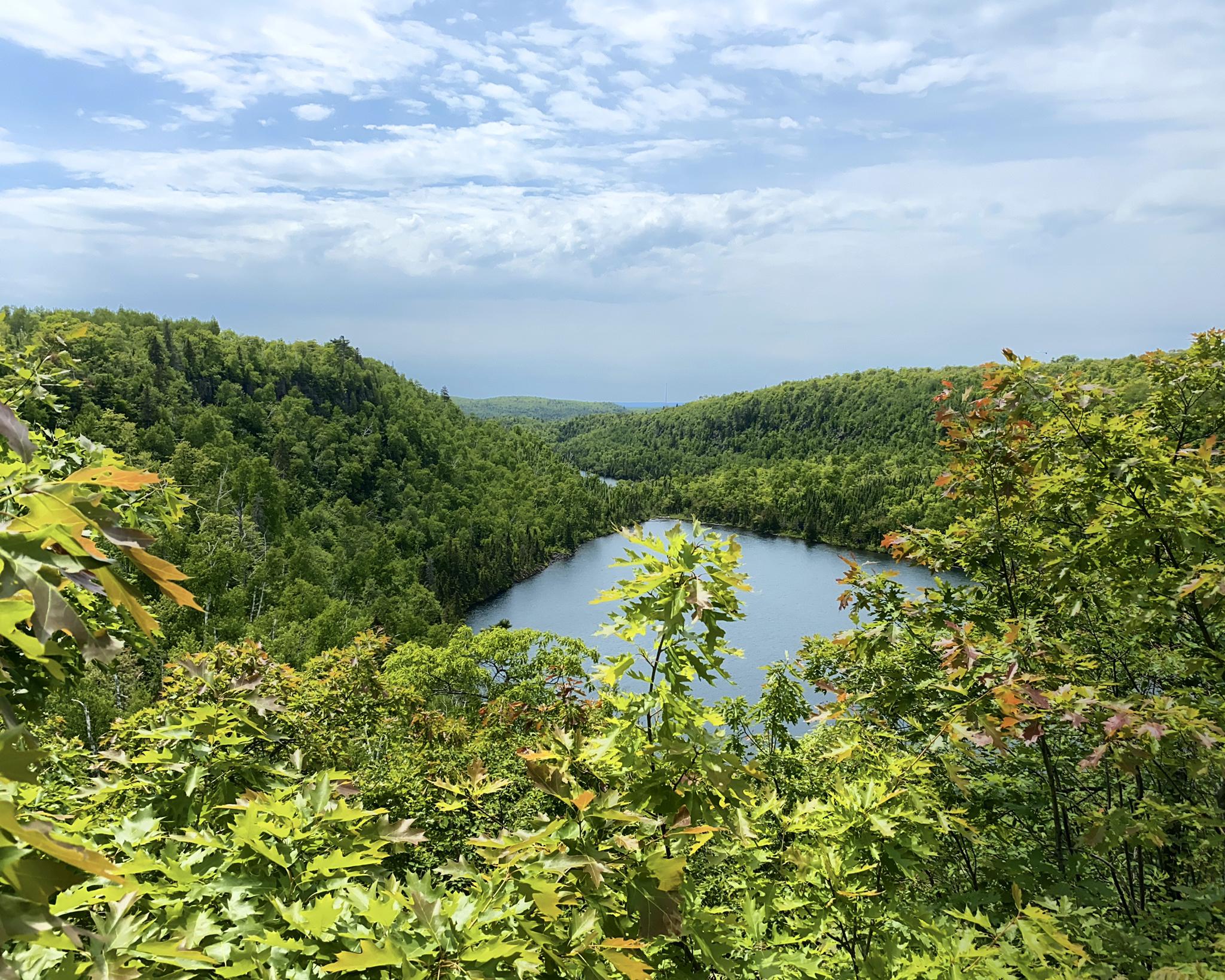 Lake nestled in dense green forest under a partly cloudy sky, seen from above.