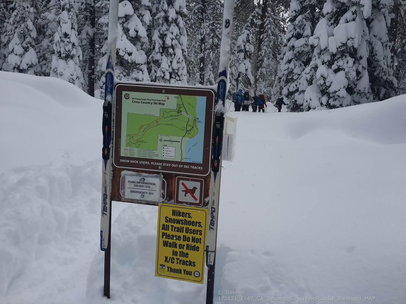 Trail sign and skis in deep snow with people in a snowy forest.