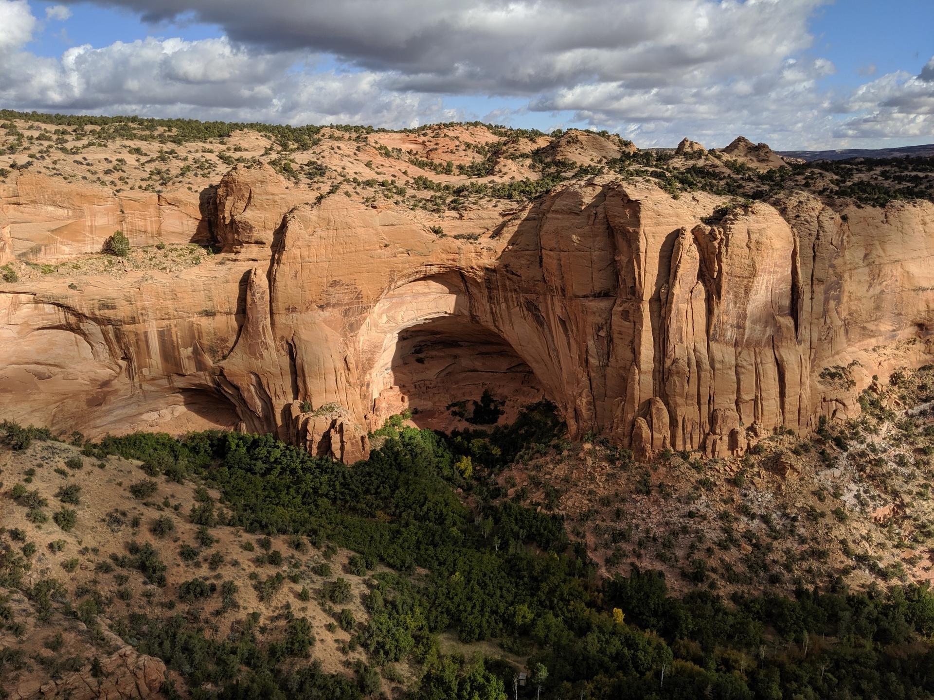 Massive natural rock arch spanning a green canyon under a cloudy sky.