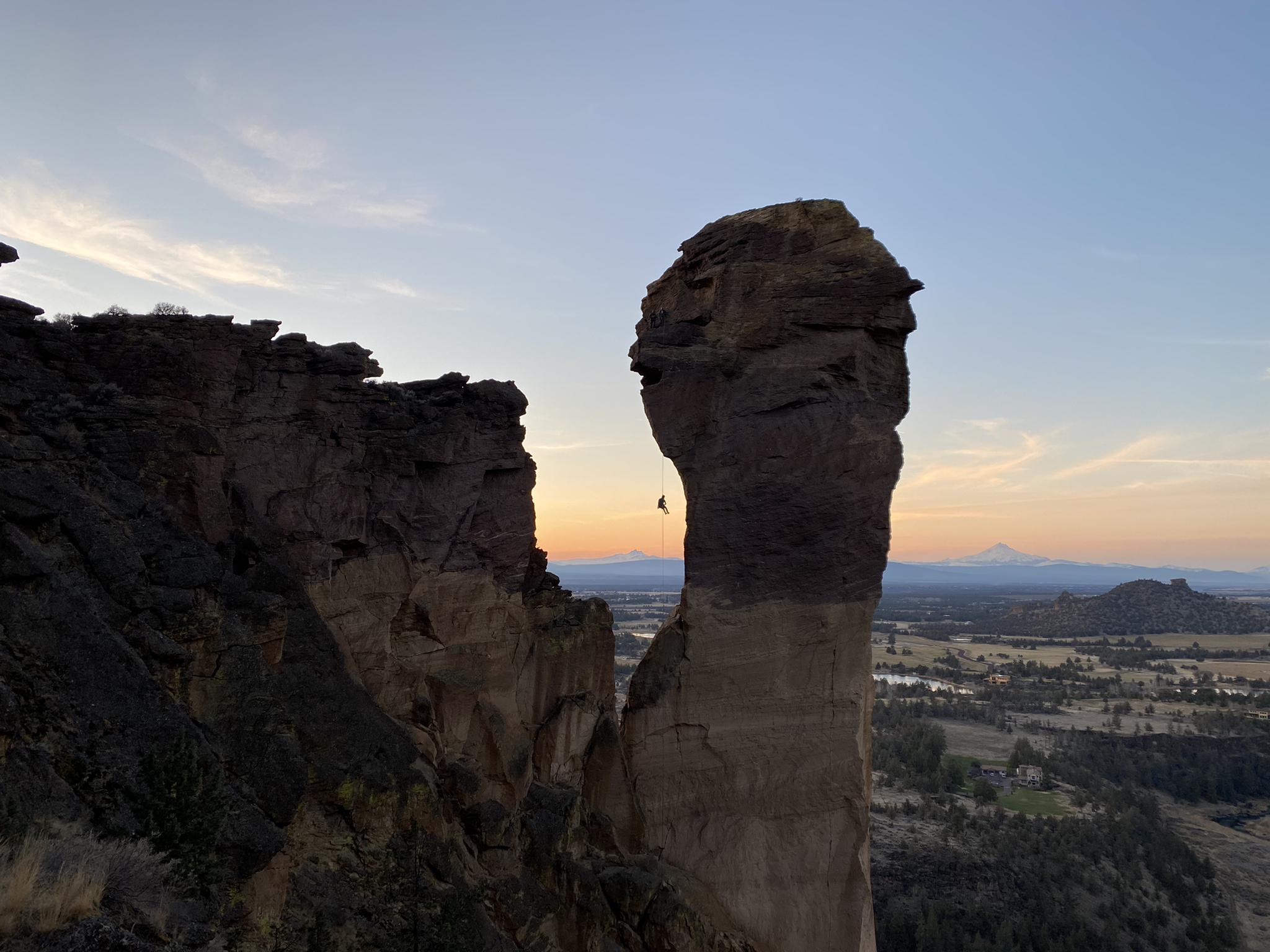A climber ascends a tall rock pillar at sunset overlooking a vast landscape.