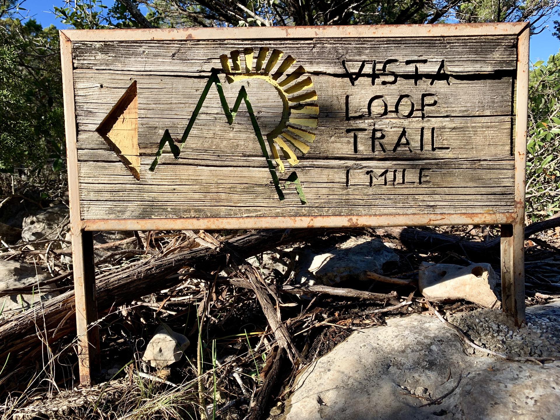 Rustic wooden sign with "Vista Loop Trail 1 Mile" carved. Sunny.
