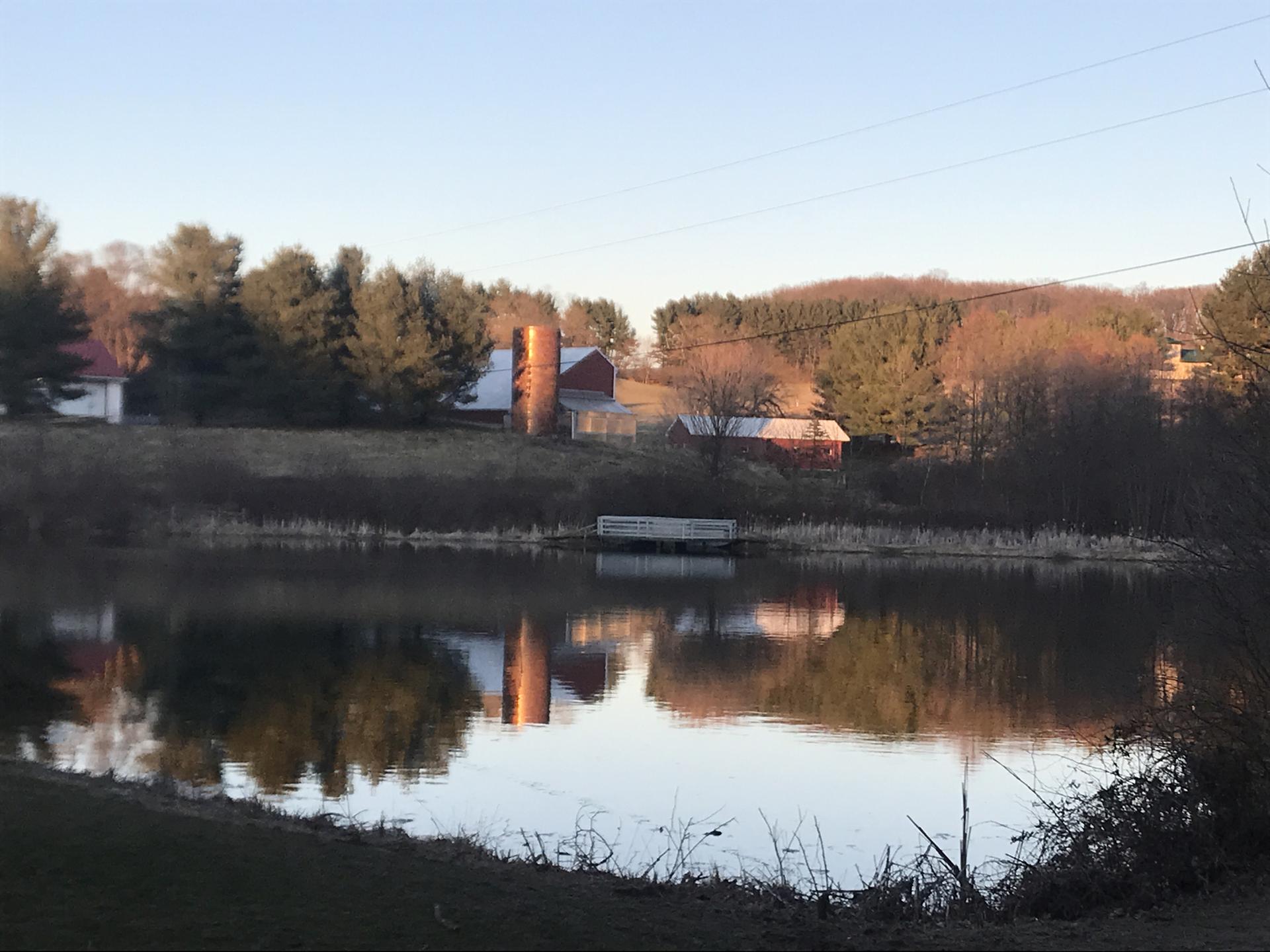 A calm pond reflects farm buildings and trees under a clear morning sky.
