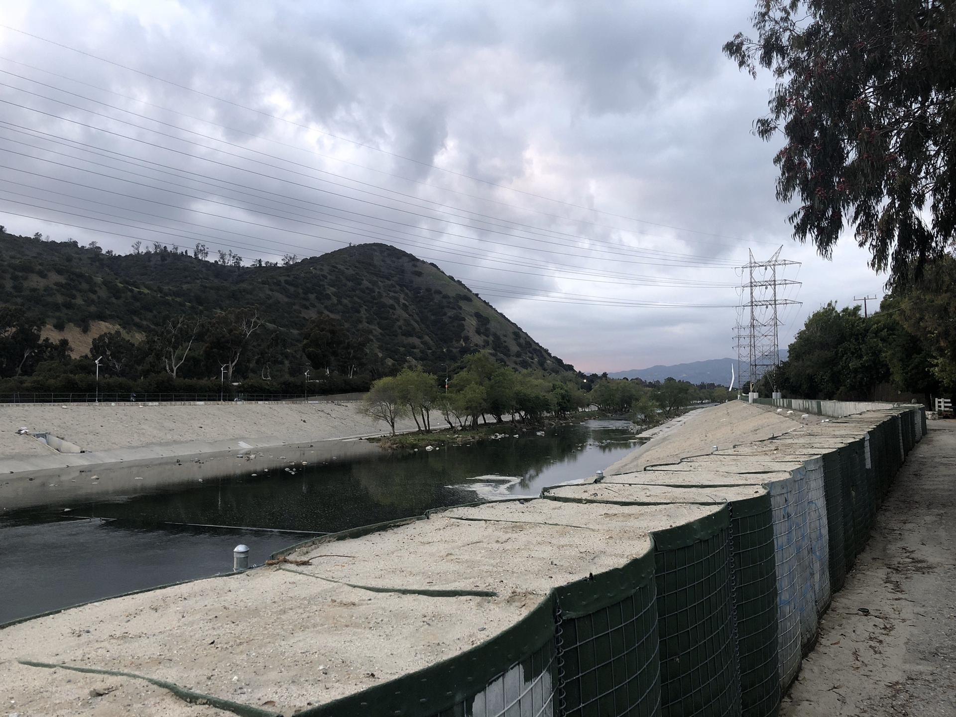River in concrete channel with dirt bank, fence, hills under cloudy sky.