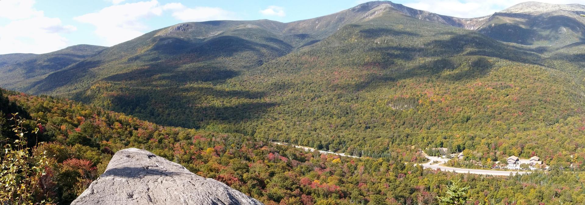 Panoramic view of vast green and autumn-colored mountains under a partly cloudy sky.