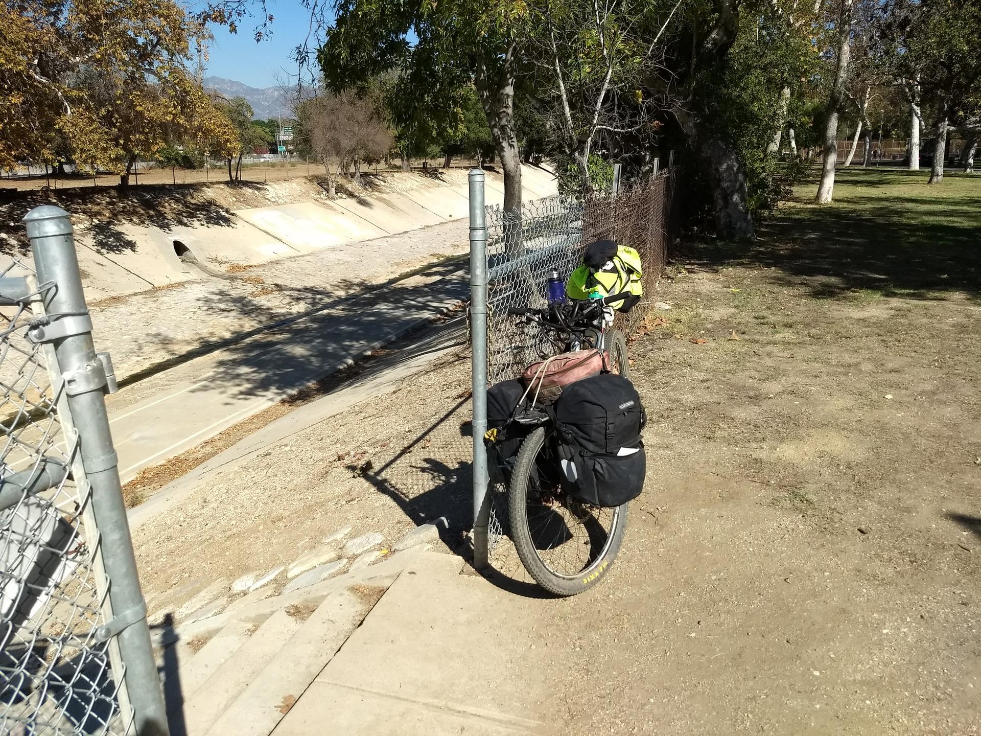 Bicycle with panniers parked by a fence next to a dry canal.