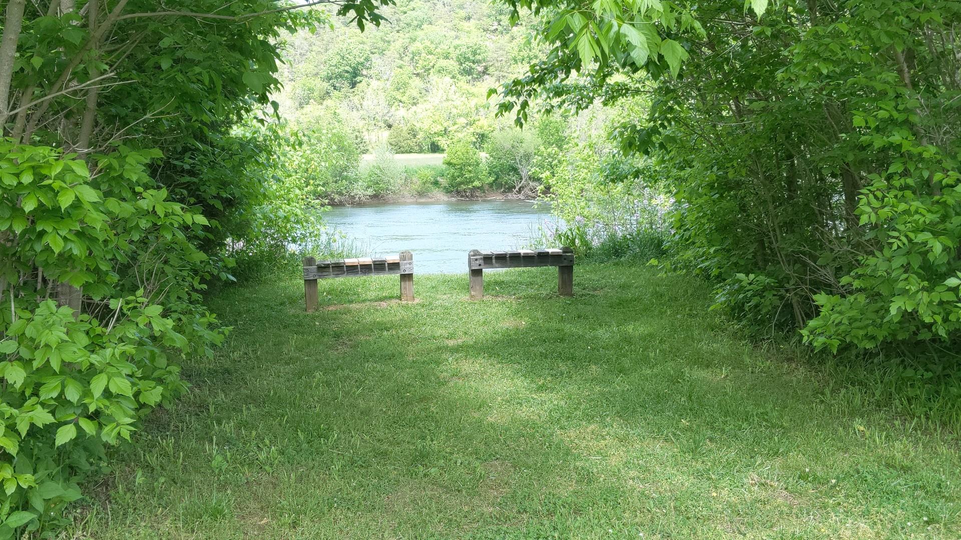 Two wooden benches overlook a calm river, framed by green trees.