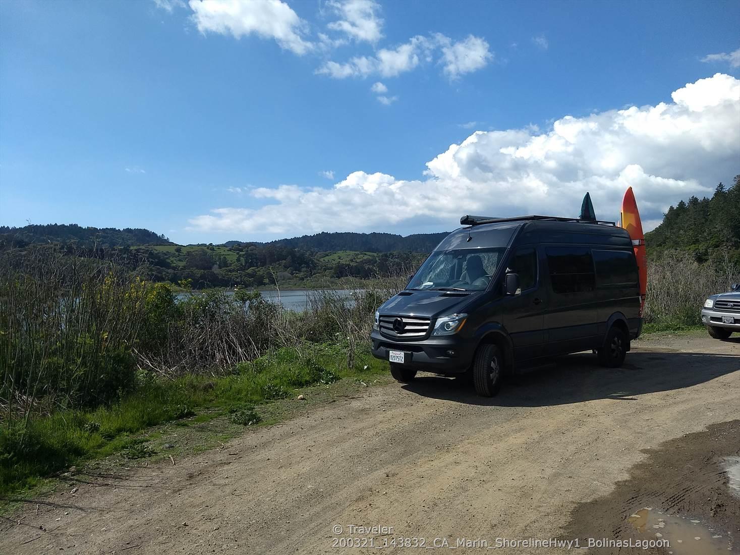 Black van with surfboards parked on a dirt road by a lake.