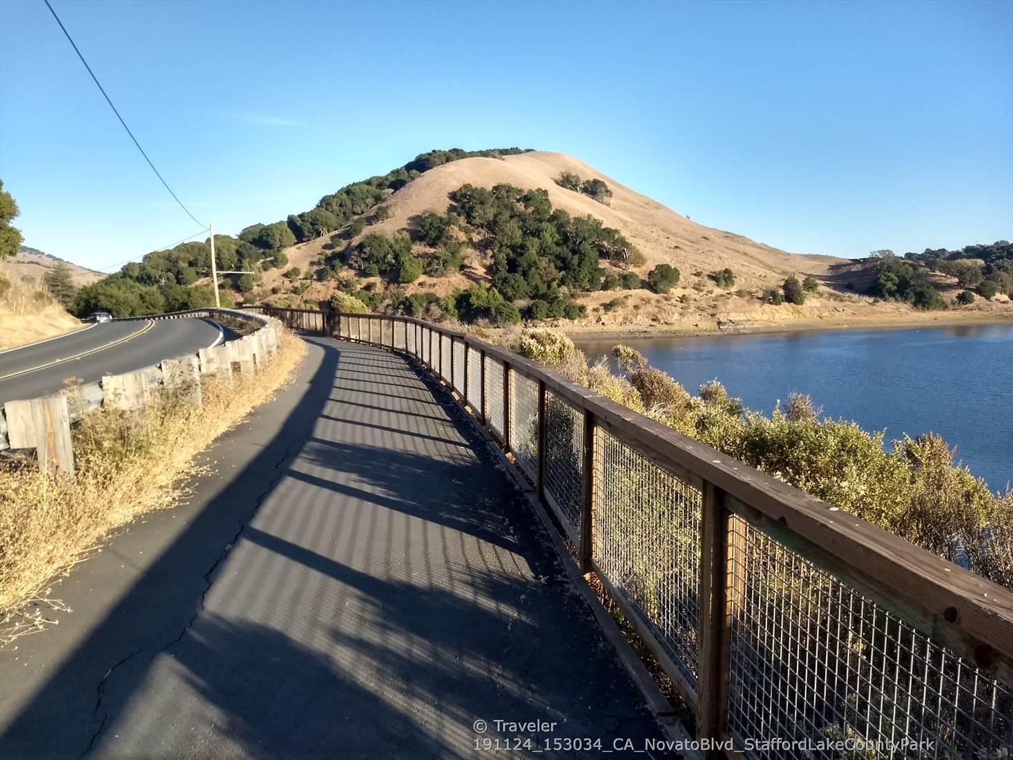 Paved path next to a lake, with dry hills and clear blue sky.