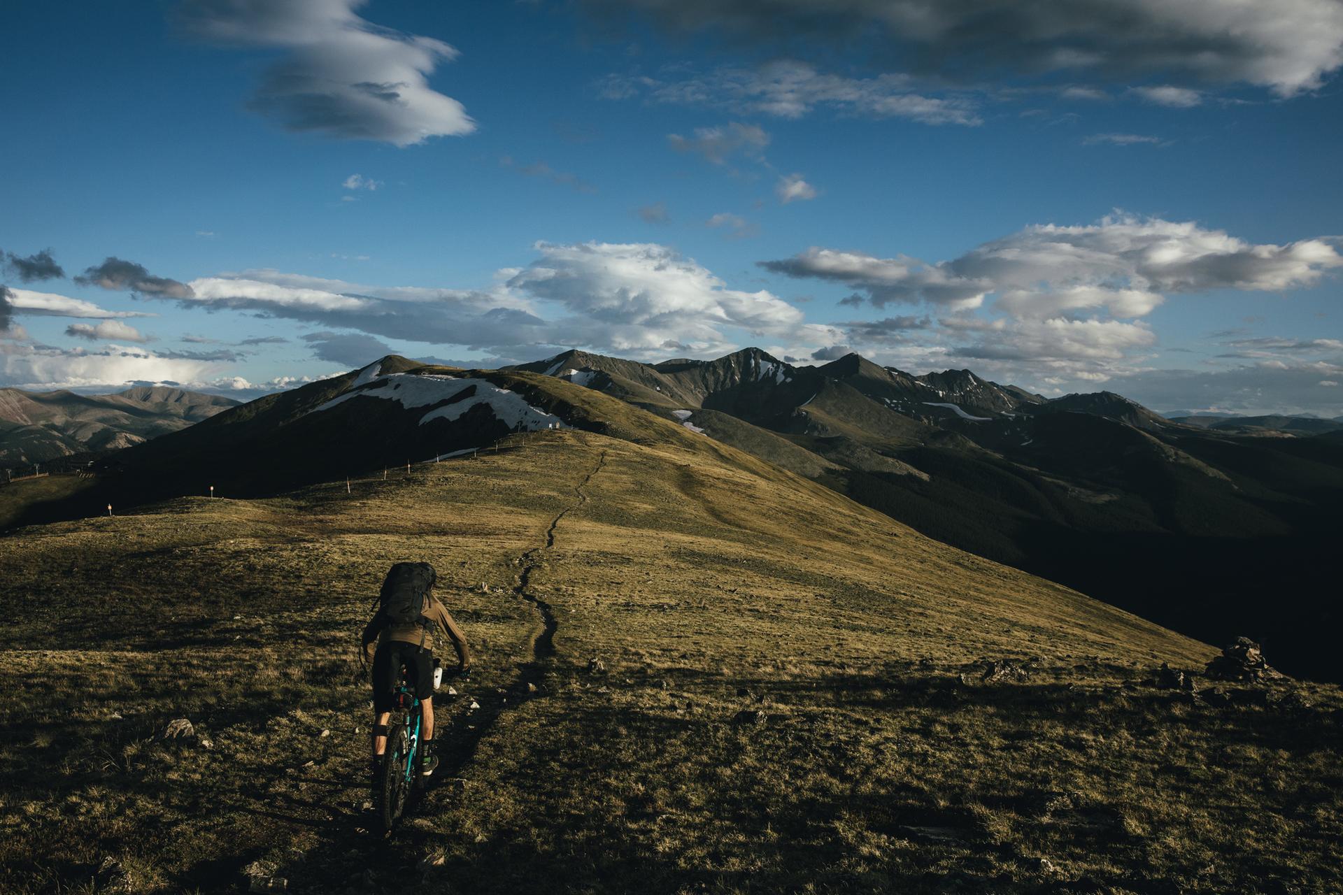 Person pushing a mountain bike up a winding path on a vast mountain.