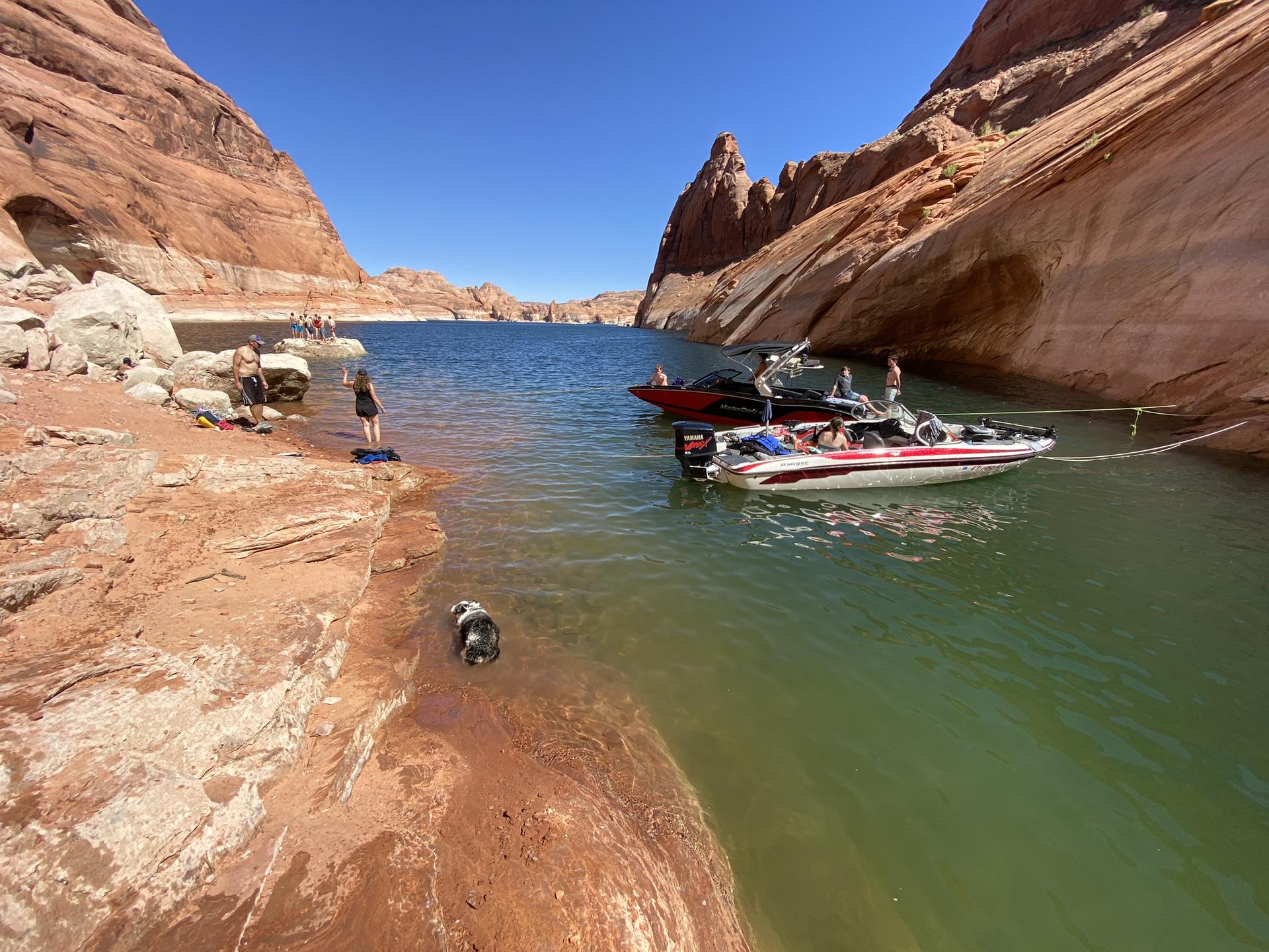 People and boats by a red rock shore in clear canyon water.