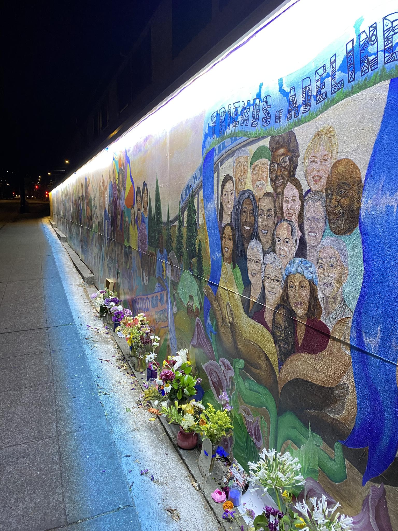 A vibrant mural depicting diverse faces, illuminated at night with flowers below.