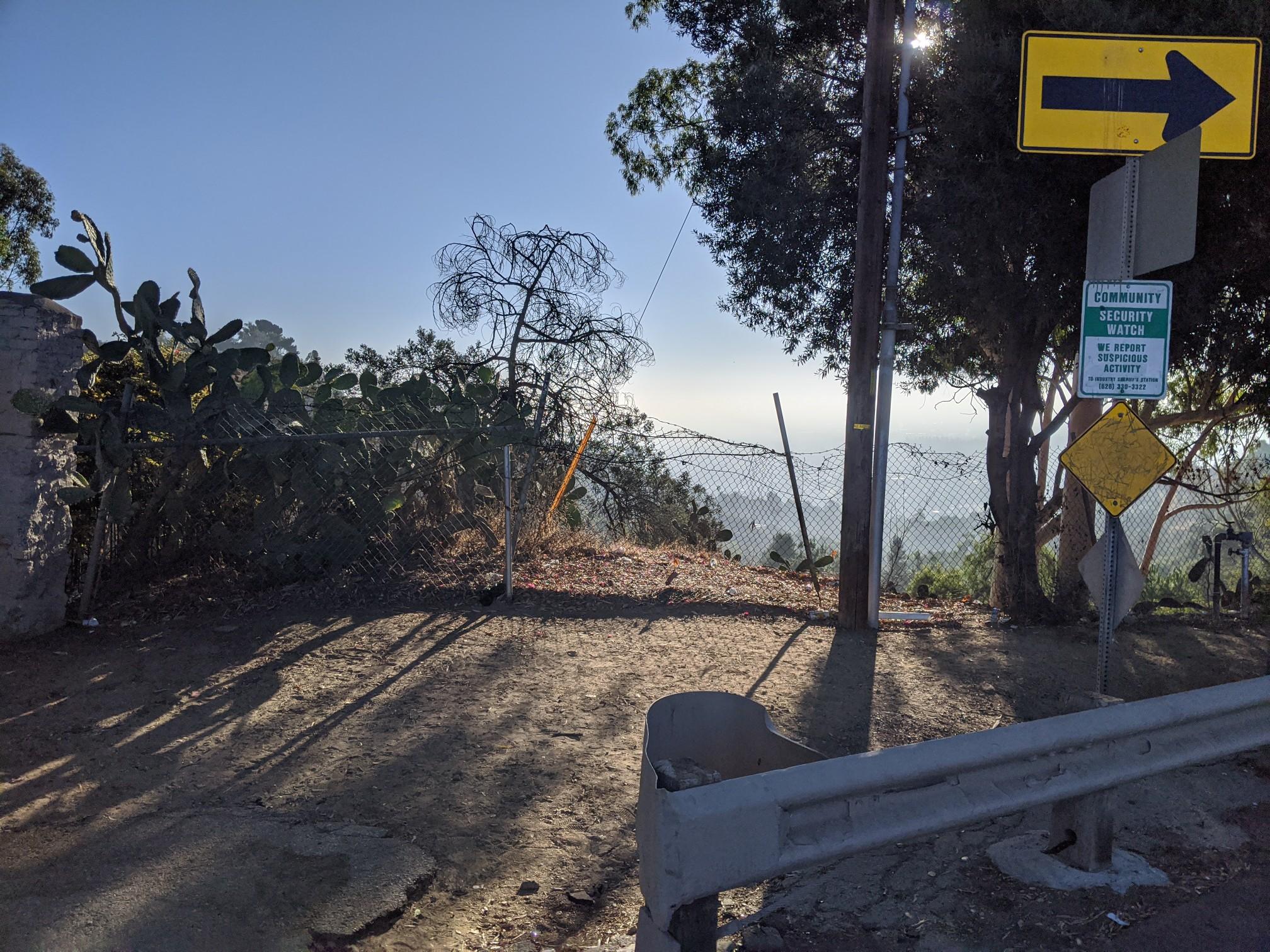 Sunny dirt path overlooking a hazy distant city, with signs and trees.