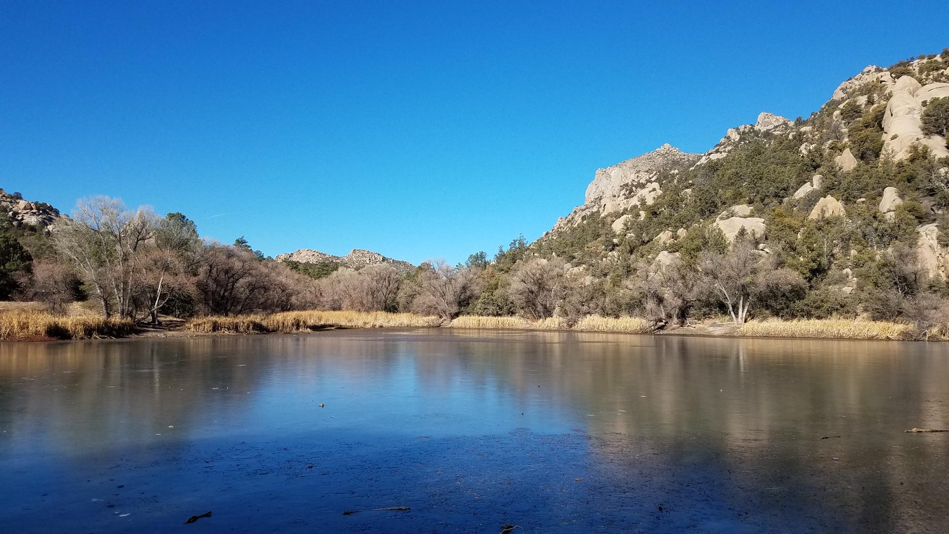 Frozen lake surrounded by bare trees and rocky hills under clear blue sky.