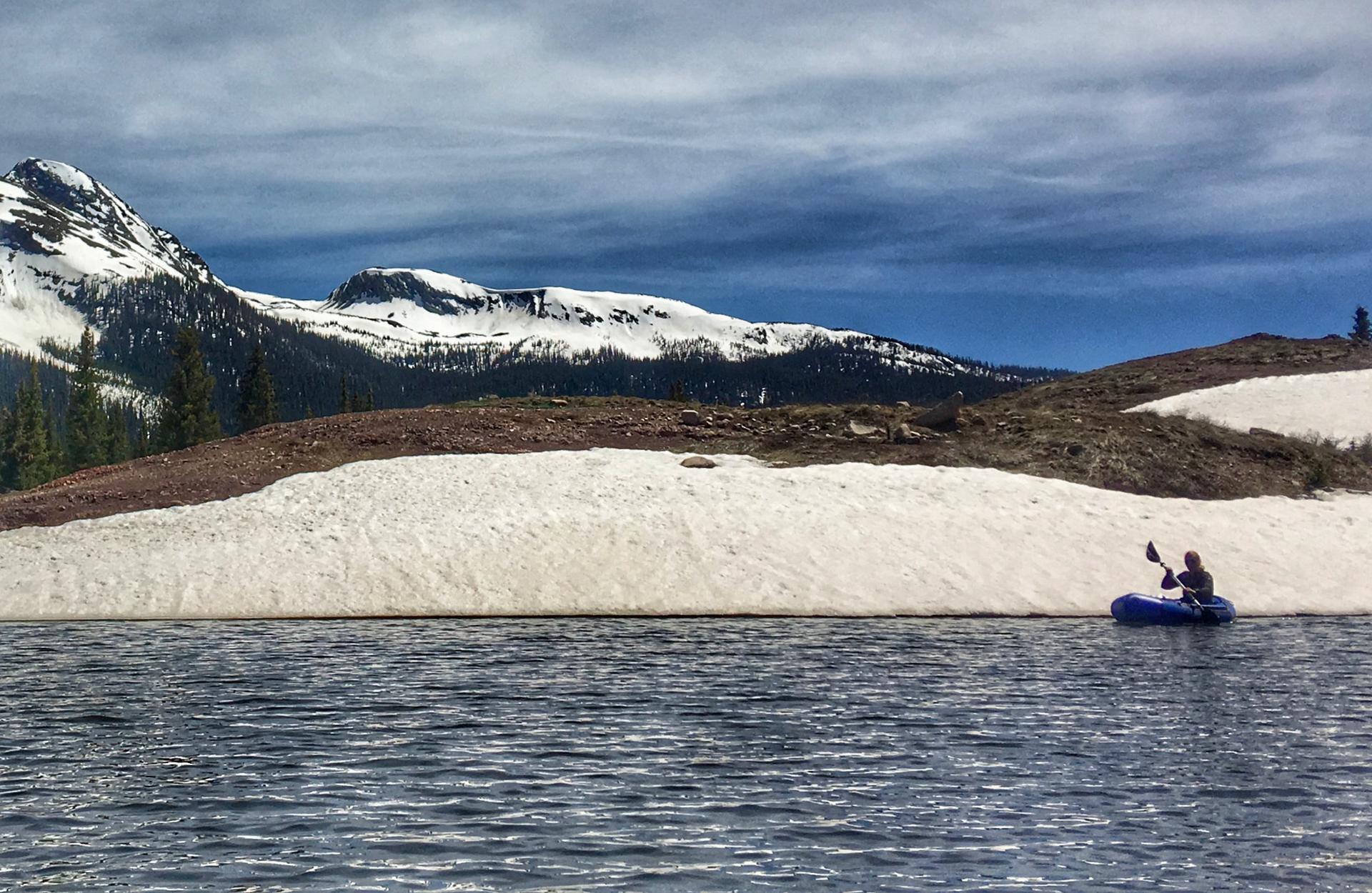 Kayaker on a lake near snowy banks and mountains under a bright, cloudy sky.