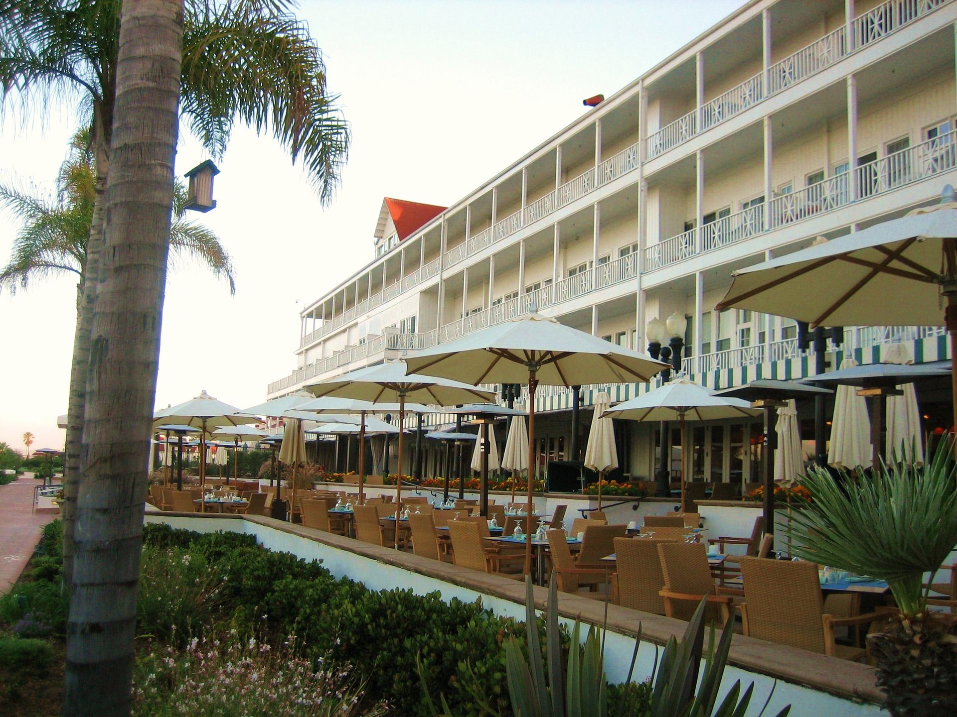 Large white building with outdoor dining, umbrellas, and palm trees under soft daylight.