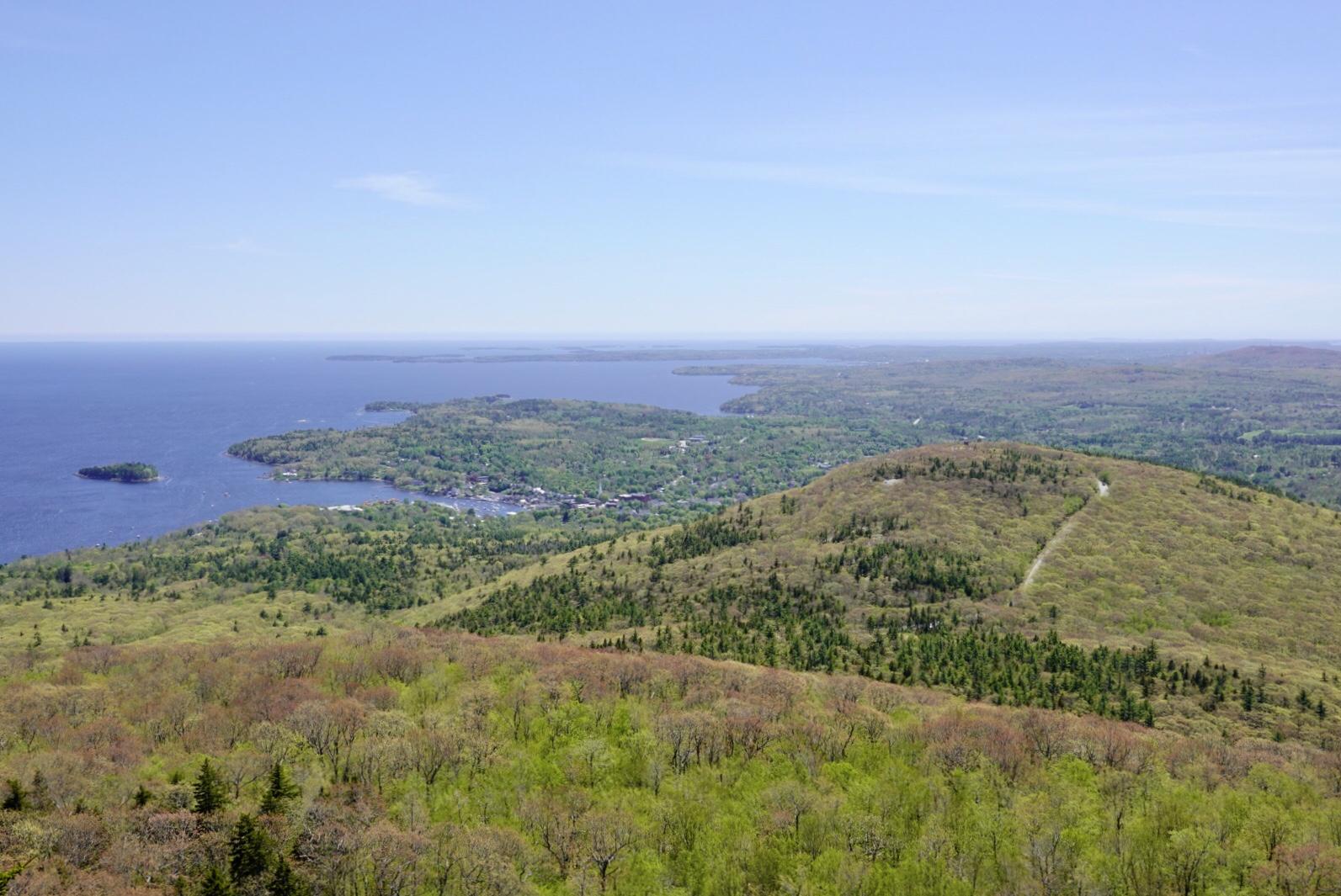 Expansive view of green forested hills, a bay, and distant ocean under clear sky.